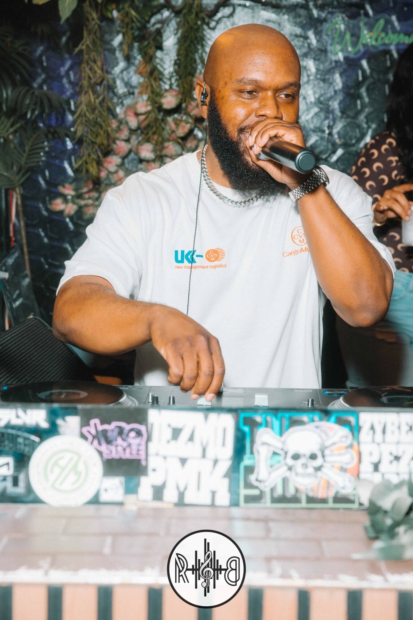 Man with a beard holding a microphone, DJ equipment in front, colorful background, wearing a white shirt.