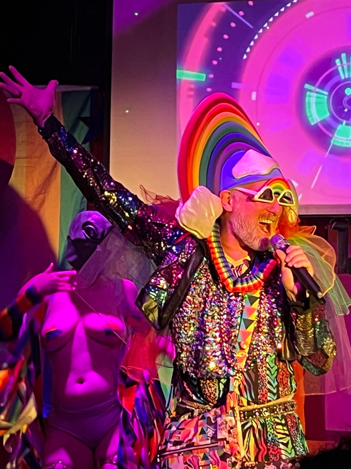 Performer dressed as a clown with rainbow hair, colorful outfit, and sunglasses, singing into a microphone on stage with vibrant background.
