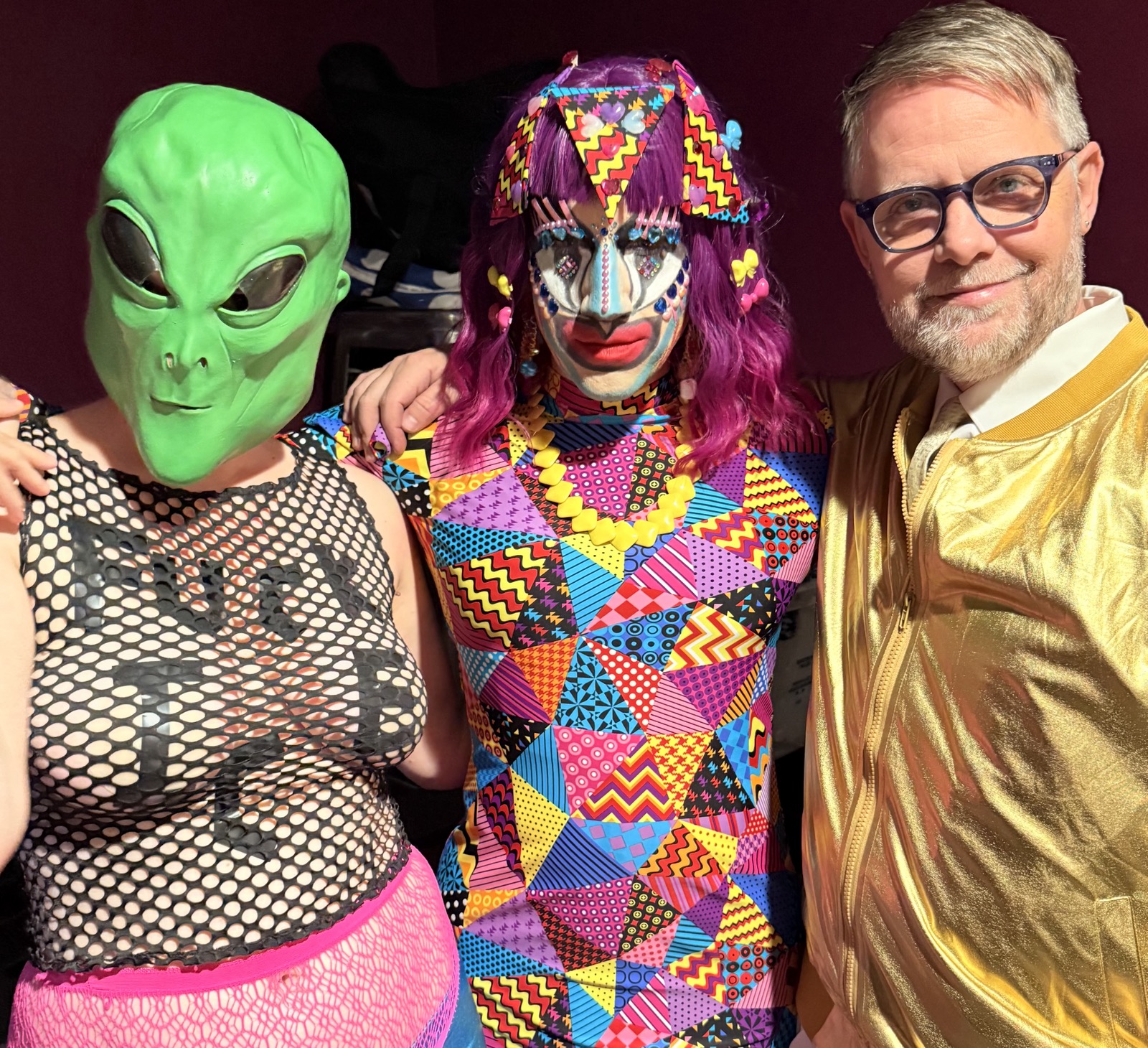 Three people in colorful costumes, one with an alien mask, one with clown makeup and vibrant clothing, and one in a shiny gold jacket.