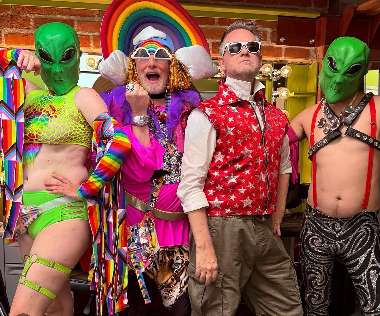 Group of four people in colorful costumes, two with green alien masks, one with rainbow headgear, and one in a red star-patterned vest, standing indoors.