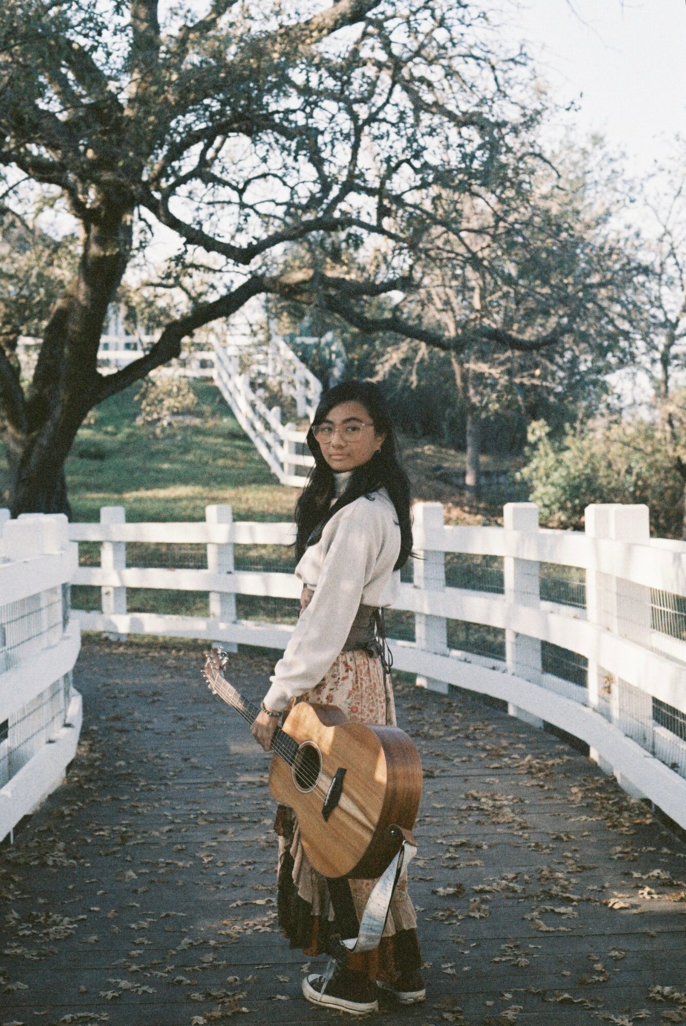 Young woman standing on a path with a guitar, white fence, trees, and stairs in the background.