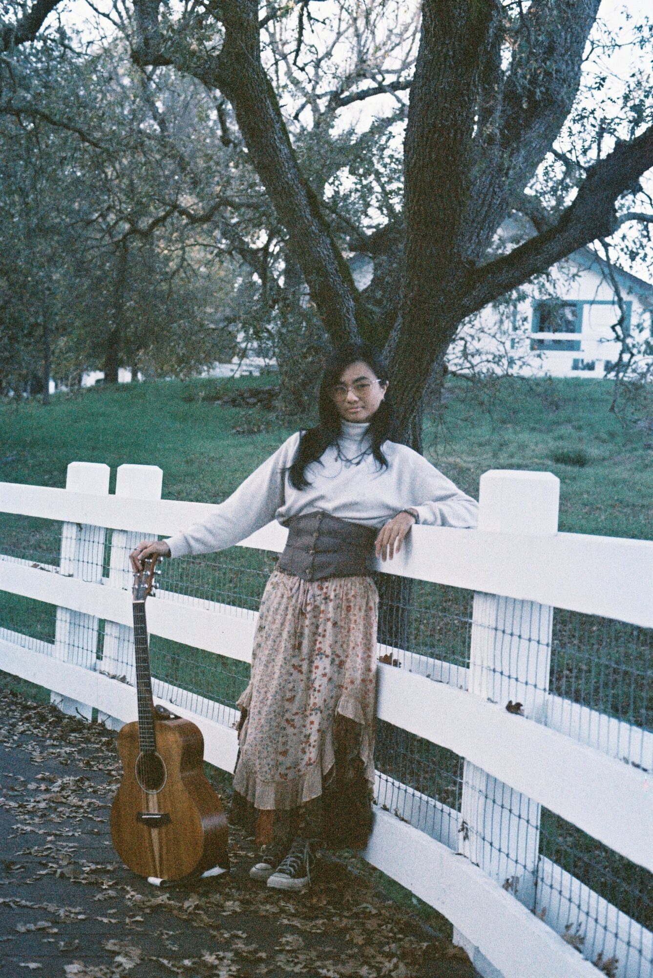 Young woman standing by a white fence with a guitar, outdoors near a large tree, smiling, daytime.