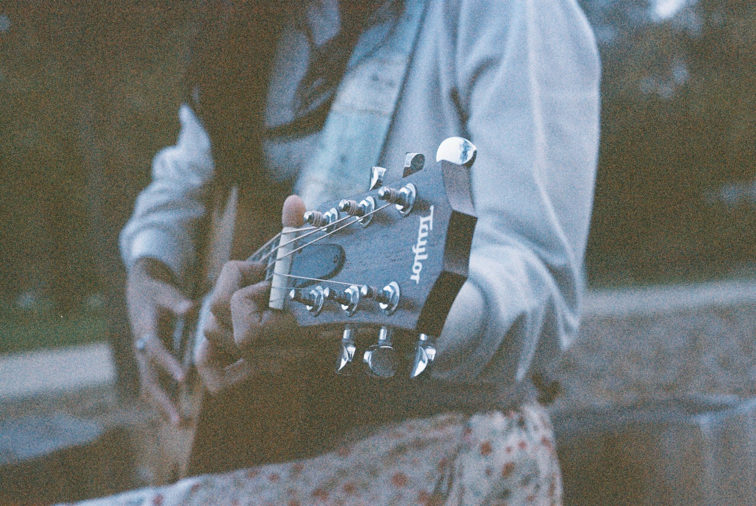 Person playing guitar outdoors, wearing a light jacket and floral skirt, with trees in the background.