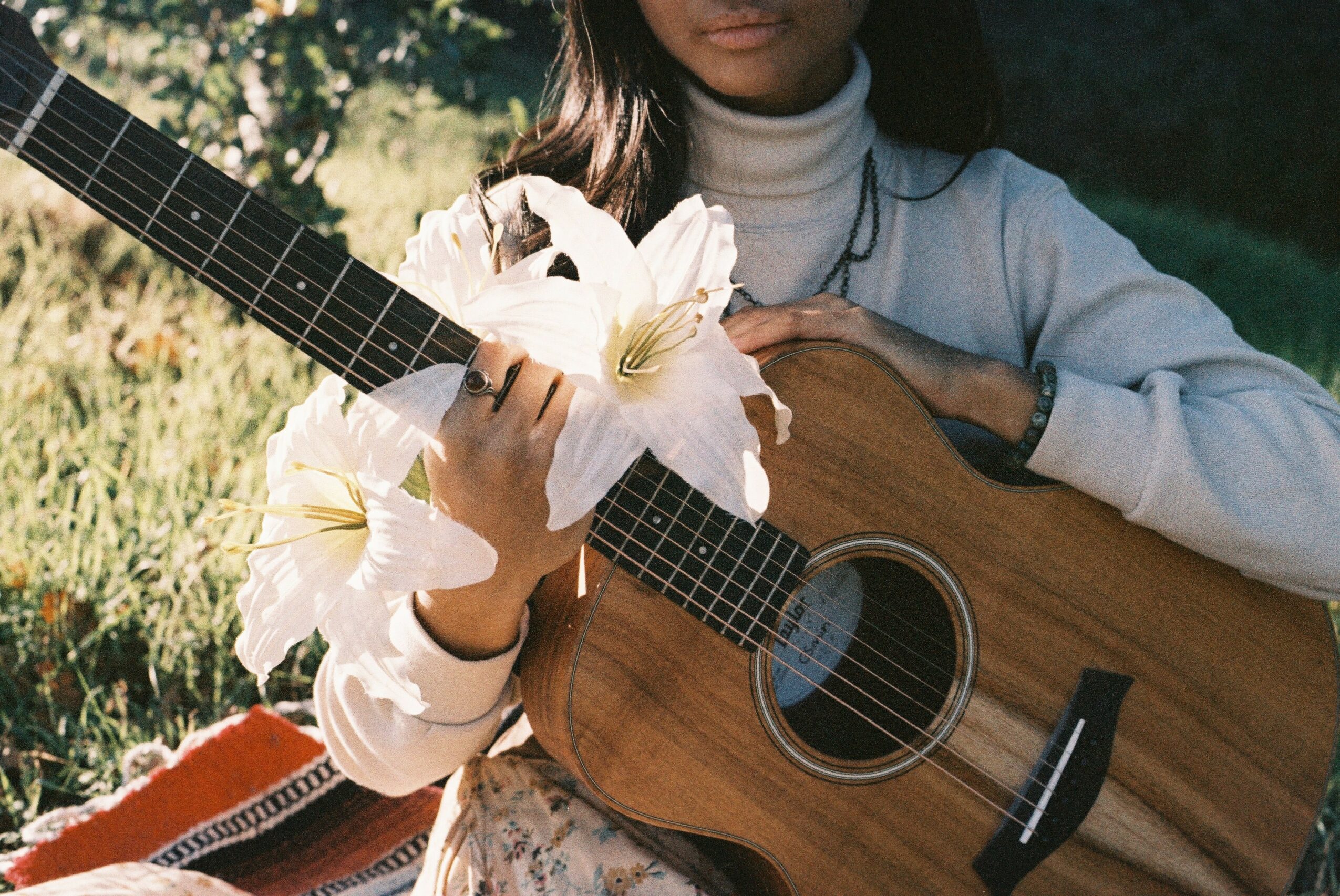 Person holding an acoustic guitar outdoors, with a large white flower near the guitar's neck, wearing a white turtleneck.
