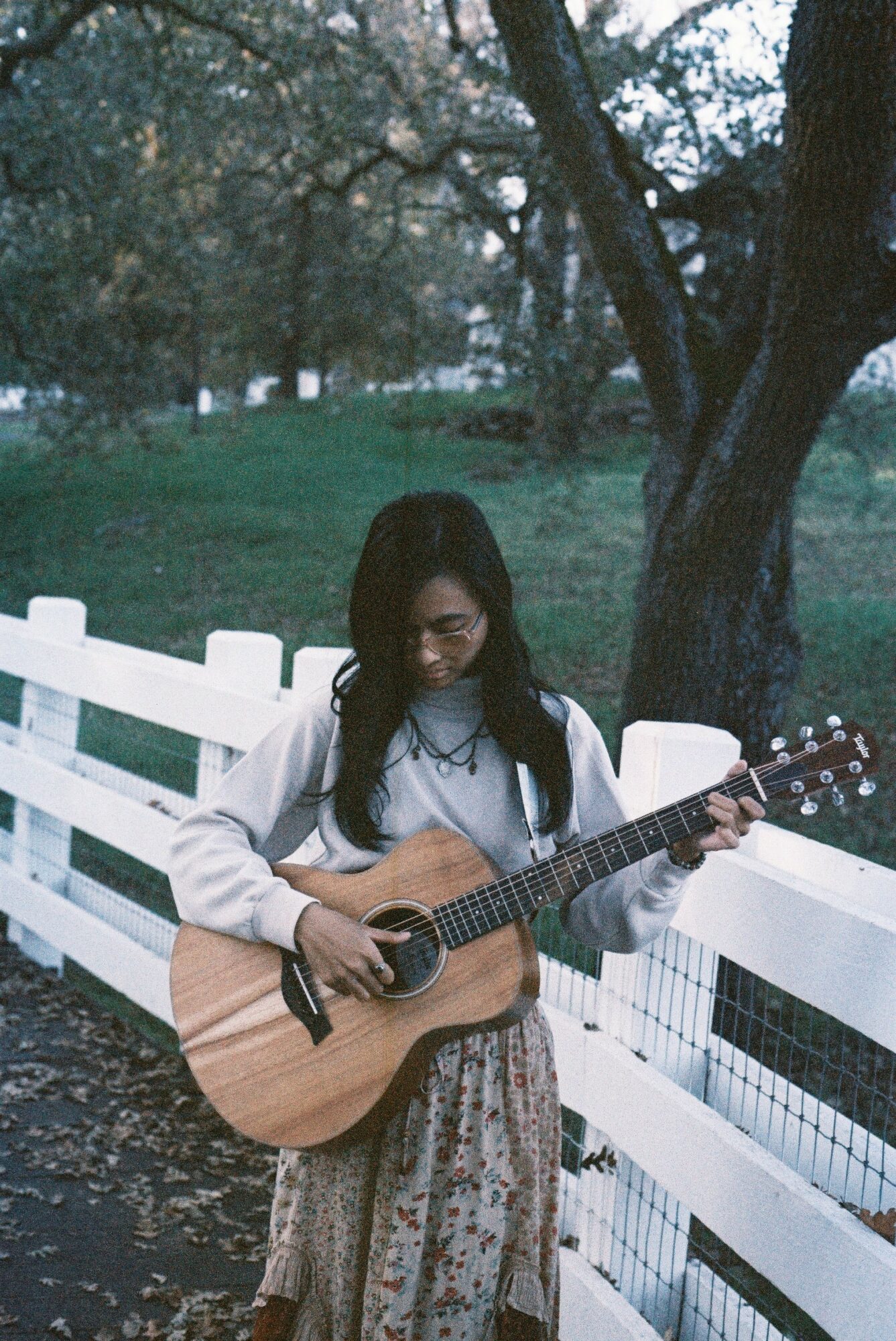 Young woman with glasses playing an acoustic guitar outdoors near a white fence, trees in background.