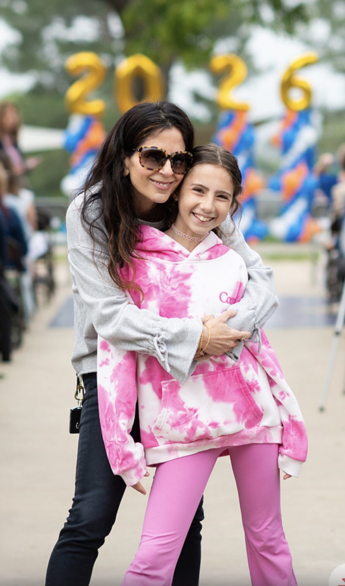 Two smiling women, one older and one younger, hugging outdoors with colorful decorations and numbers in the background.