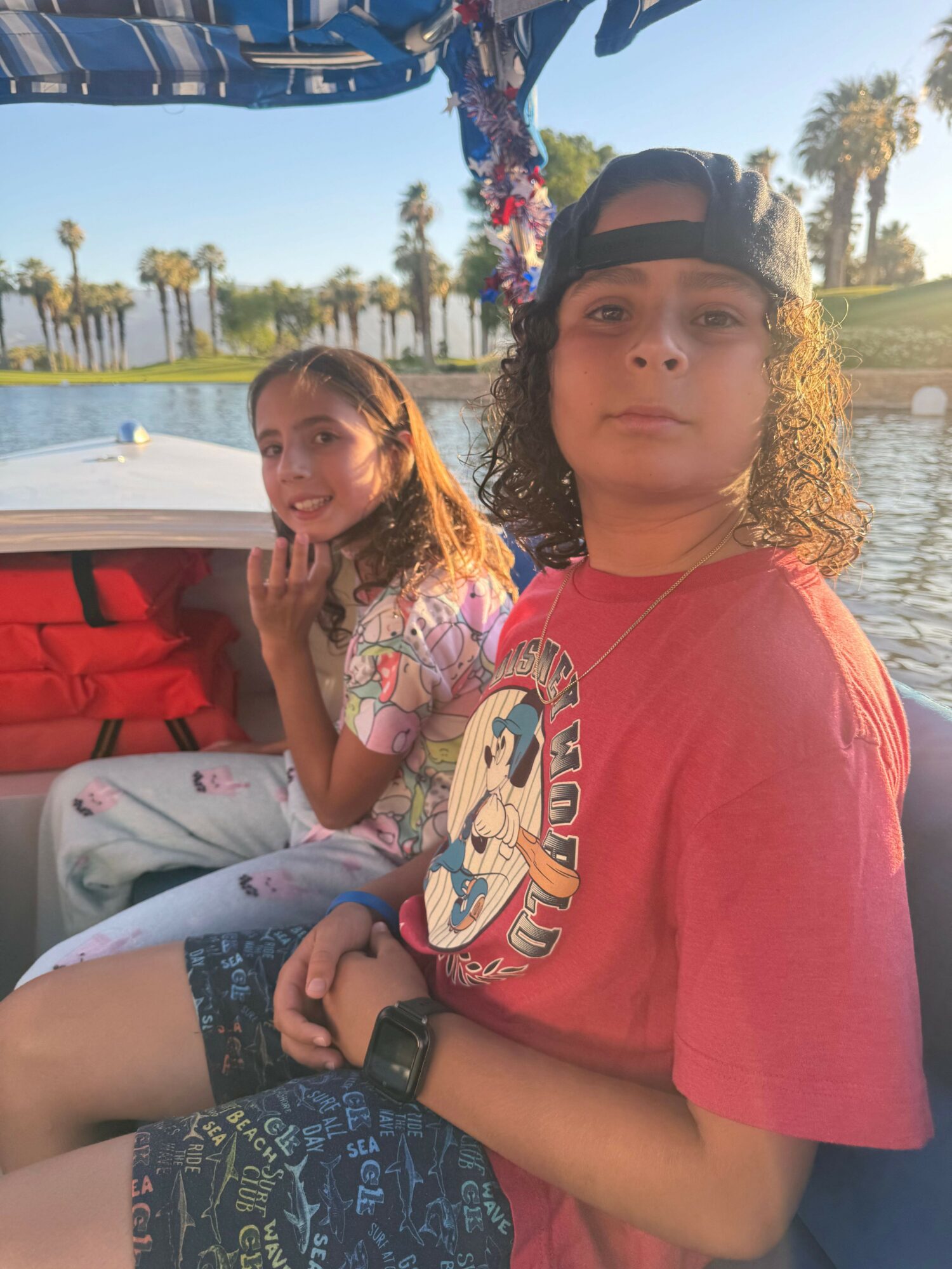 Two young women sitting outdoors near a body of water with trees and boats in the background.