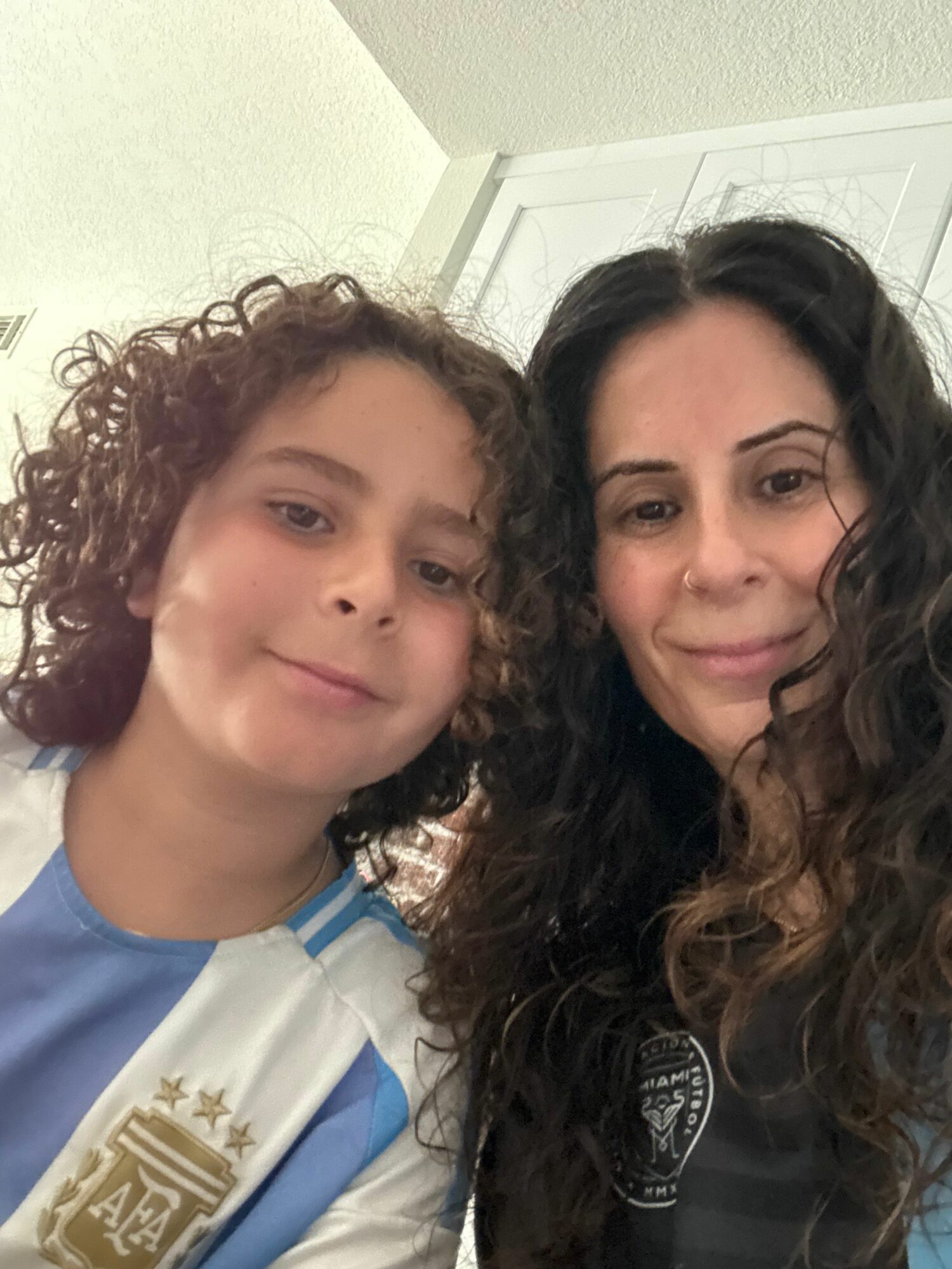 Two people smiling, one with long dark hair and the other with curly hair, indoors with a white wall background.