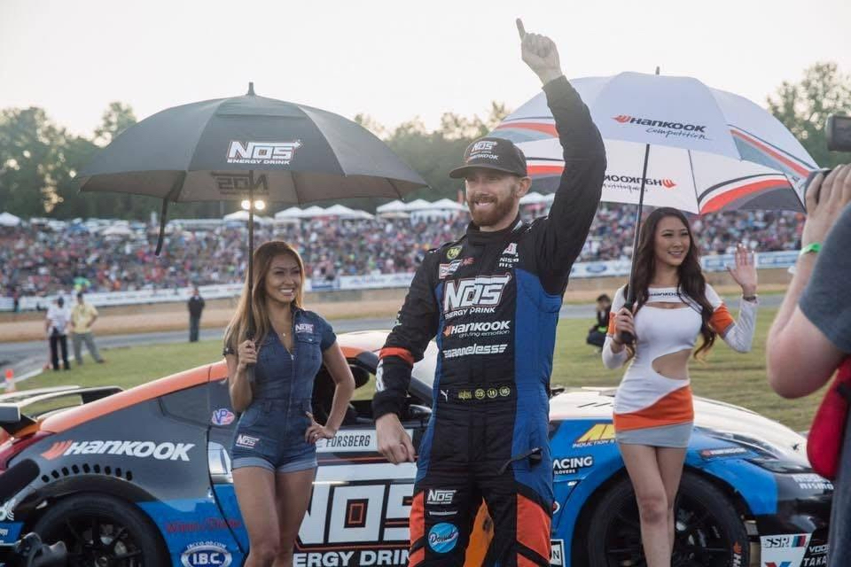 Race car driver celebrating with two women holding umbrellas at a racing event, with a crowd in the background.