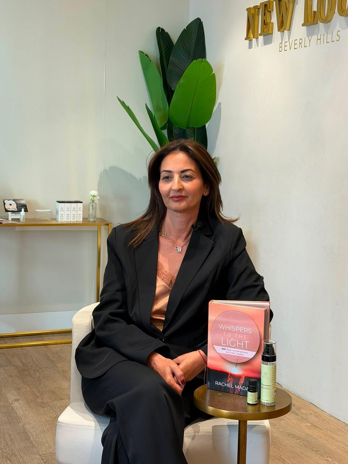 Woman in black blazer sitting at a table with skincare products, large plant, and sign in background.
