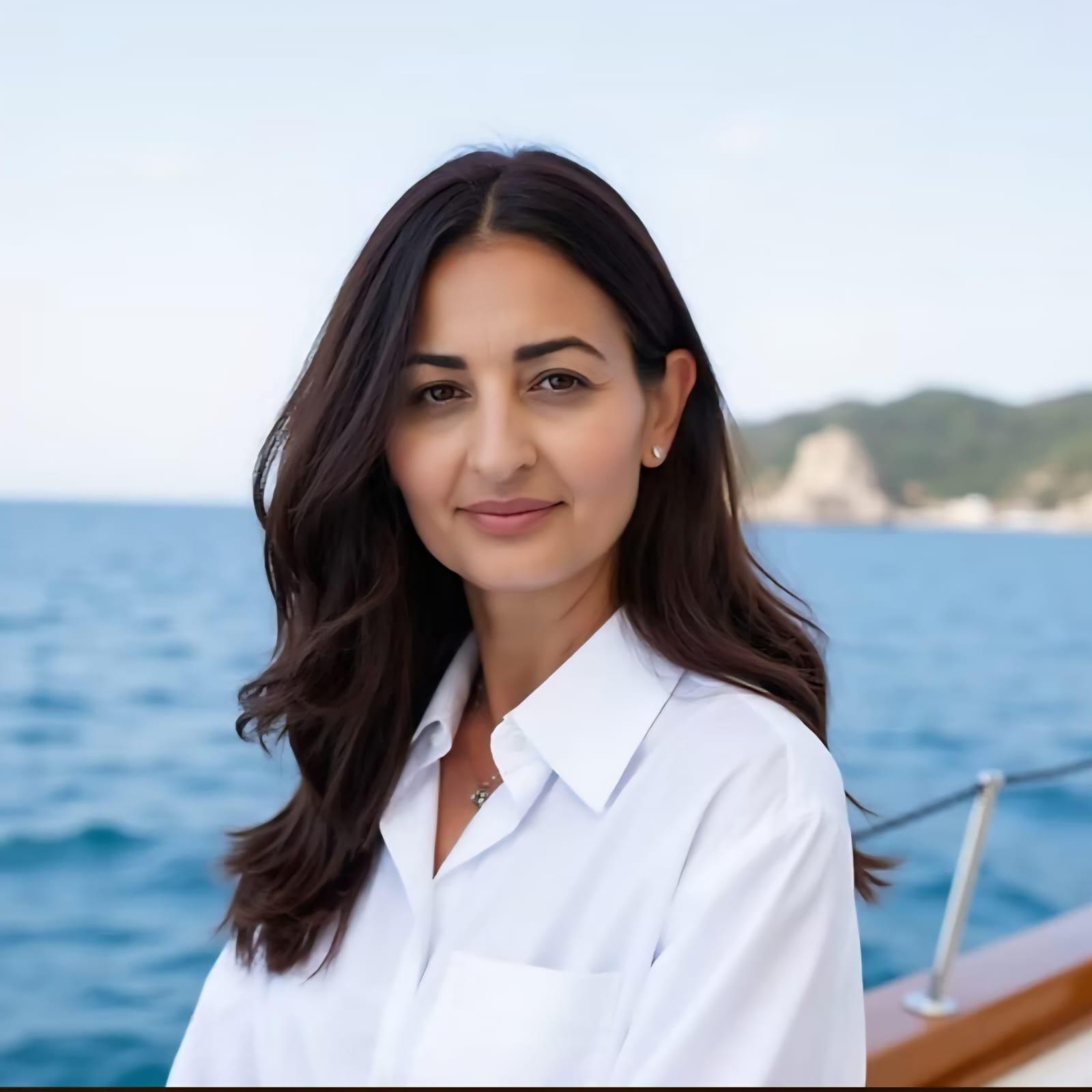 Woman with long dark hair wearing a white shirt, standing outdoors near water with a distant shoreline.