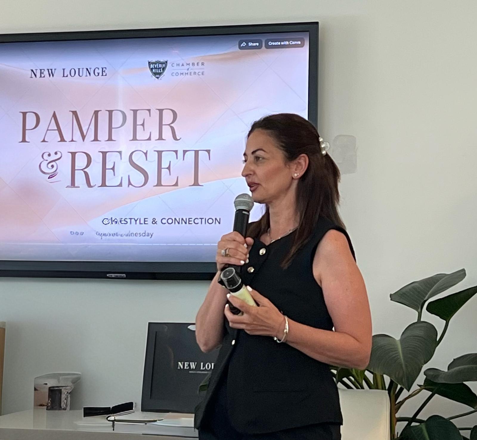 Woman speaking into a microphone in front of a presentation screen, with a plant nearby.