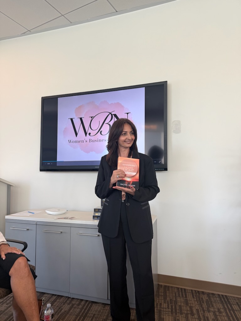 Woman in black suit holding a book, standing in front of a screen with a logo, in a conference room.