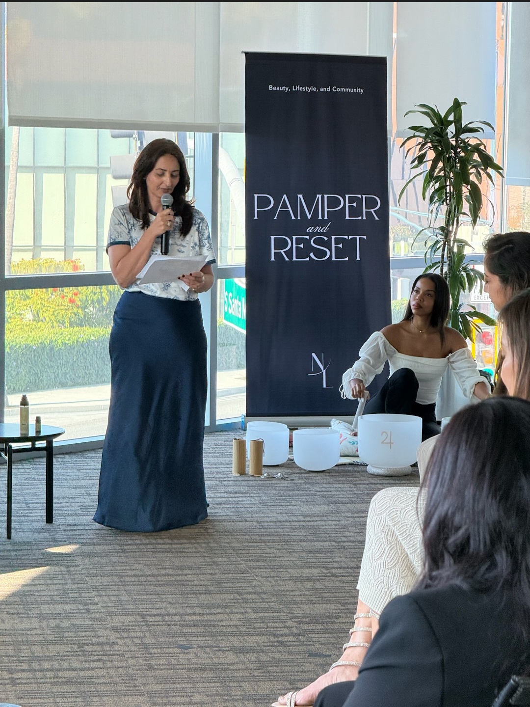 Woman speaking into microphone at event with seated audience, large window, and banner reading 'Pamper and Reset'.