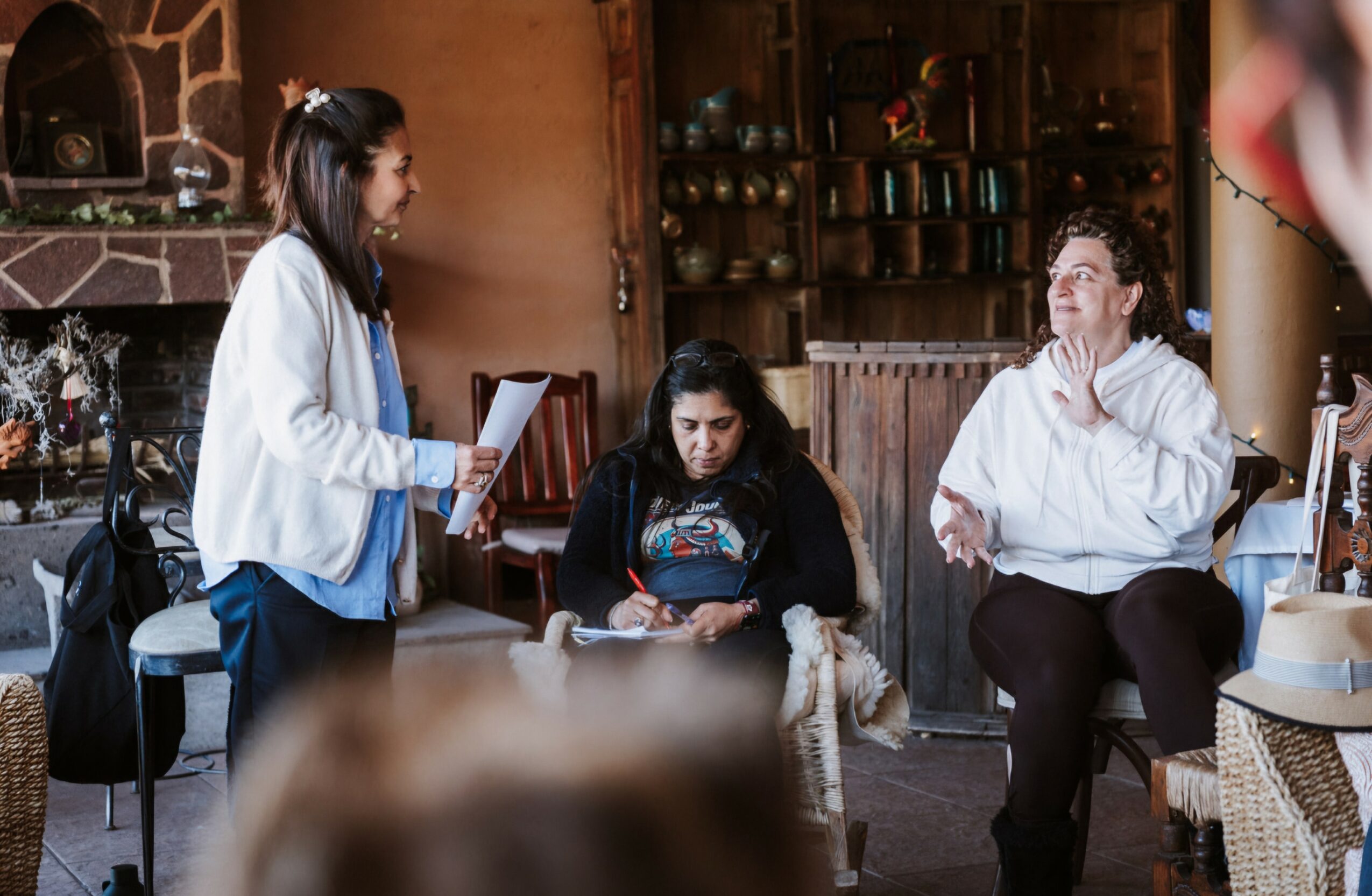 Three people in a cozy room, one standing and two sitting, engaged in conversation, with shelves and a fireplace in background.