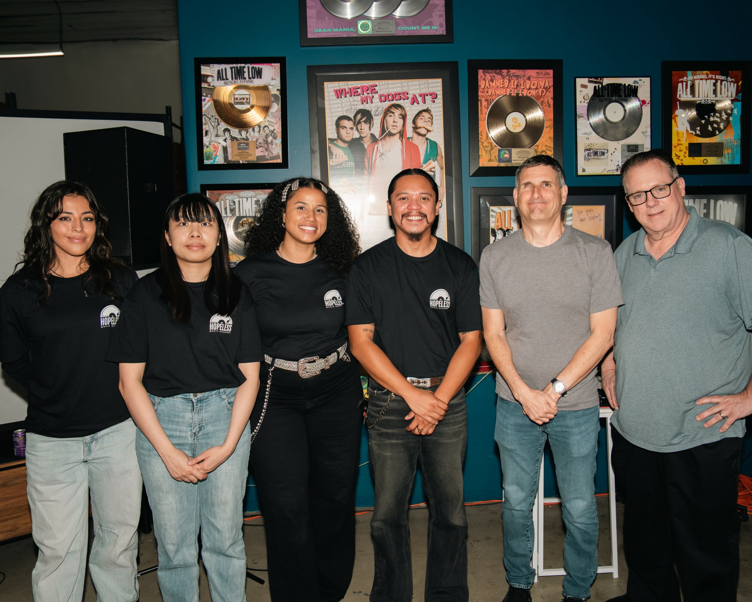 Six people standing in front of a wall with framed music awards and posters, smiling at the camera.
