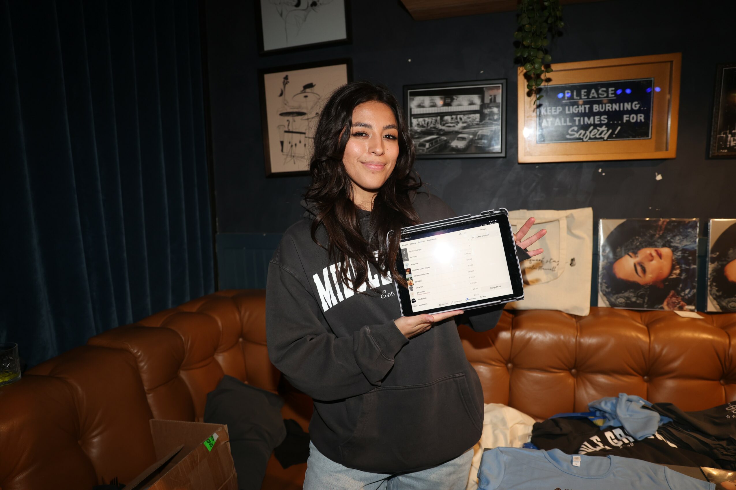 Young woman with dark hair holding a tablet, sitting on a brown leather couch in a room with framed pictures and posters on the wall.