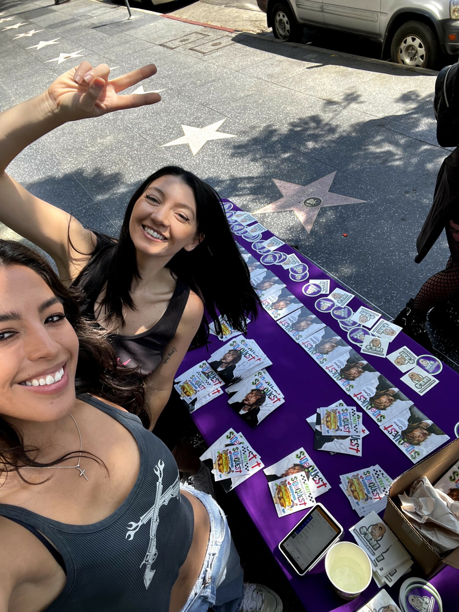 Two women smiling at a table with cards and papers outdoors, one making a peace sign, shadows and parked cars in background.