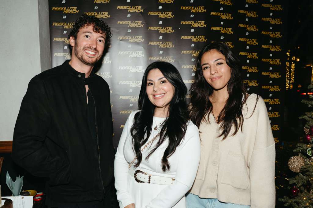Three people standing together in front of a black backdrop with gold text, smiling, indoors with a Christmas tree nearby.