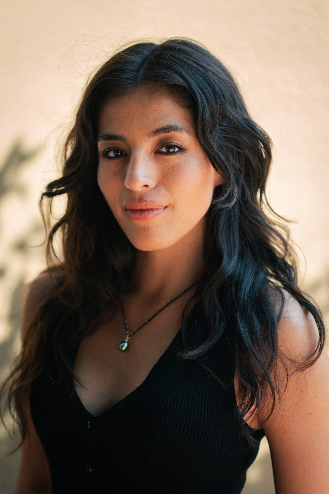 Young woman with wavy dark hair, wearing a black top and necklace, smiling softly outdoors against a neutral background.