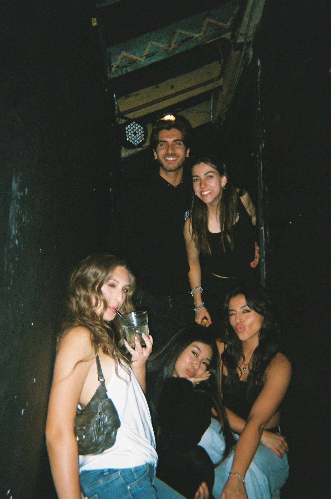 Group of five young people smiling and posing together in a dark indoor setting, some holding drinks.