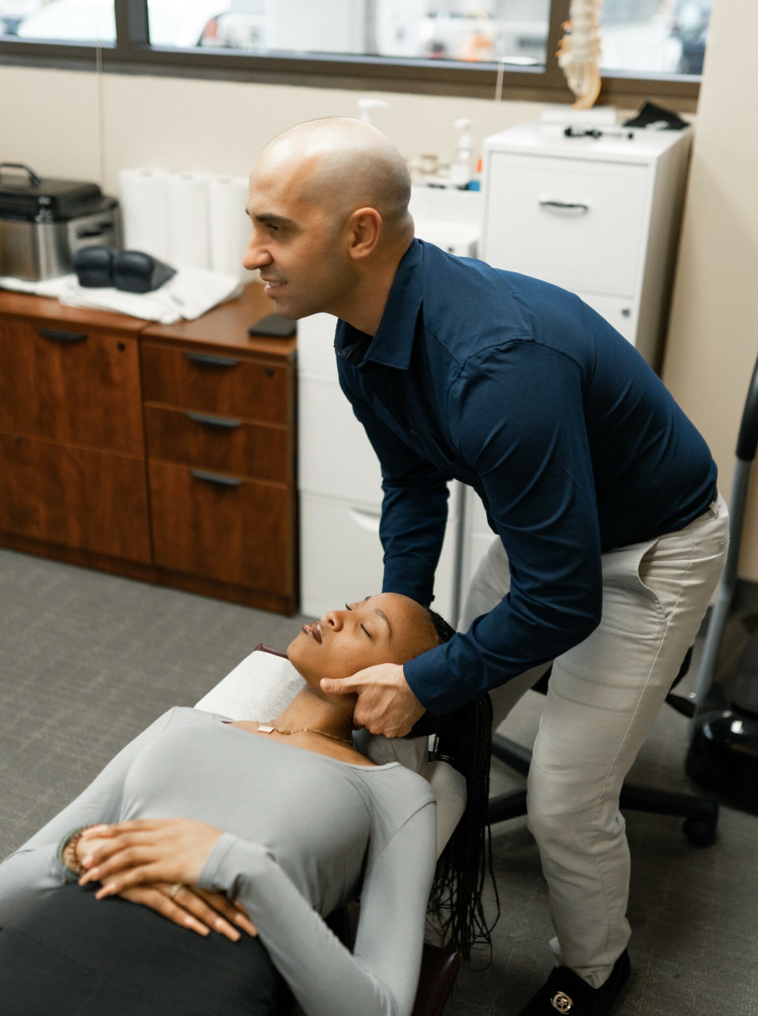 A man performs a neck adjustment on a woman lying on a treatment table in an office setting.