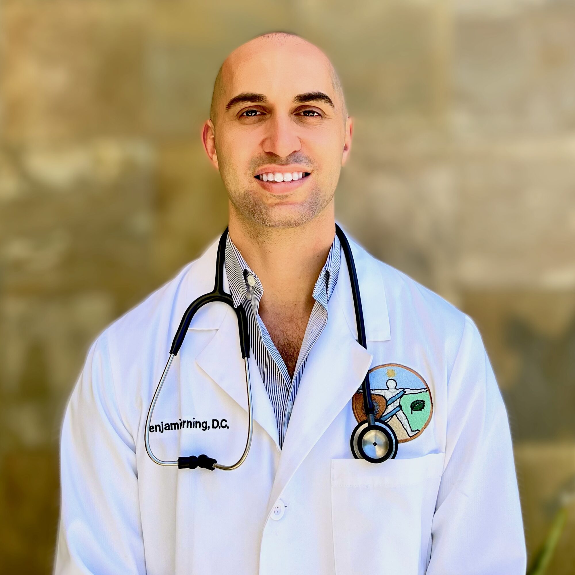 Male doctor with stethoscope smiling, wearing white coat with a badge, standing outdoors with blurred background.