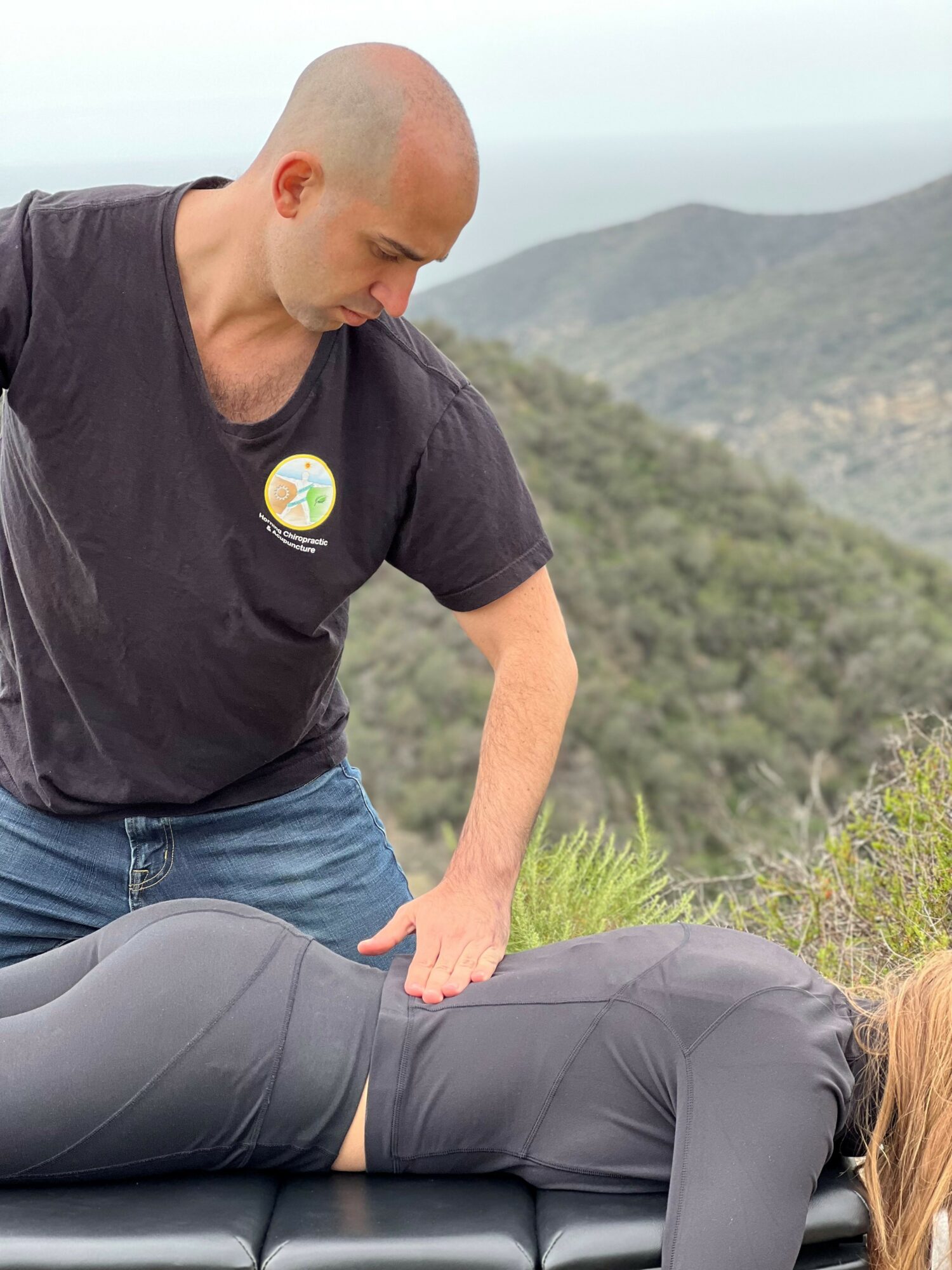 A man leans over a person lying face down outdoors with mountains in the background.