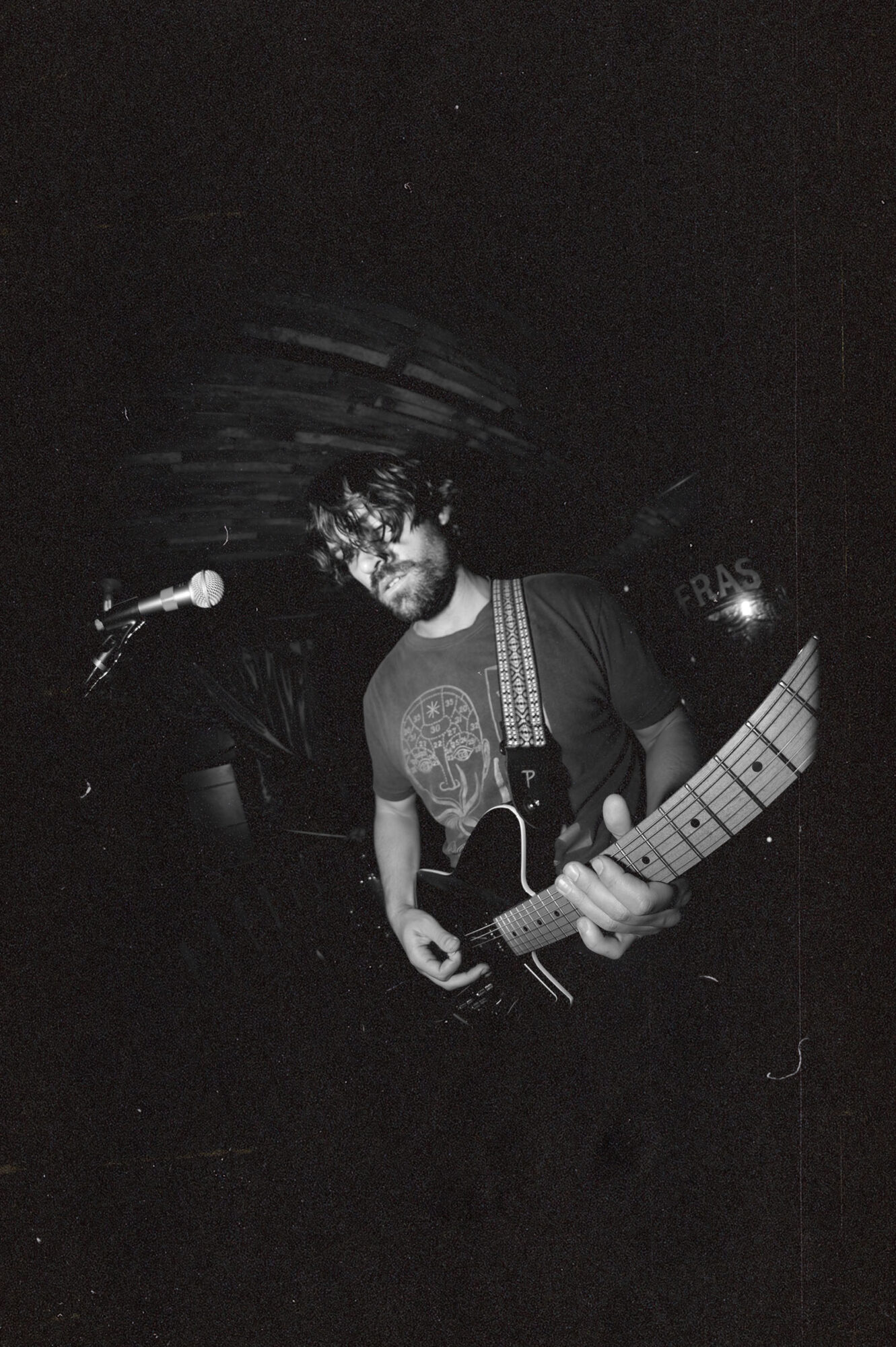 Person with curly hair and glasses playing an electric guitar on stage, dark background, microphone nearby.