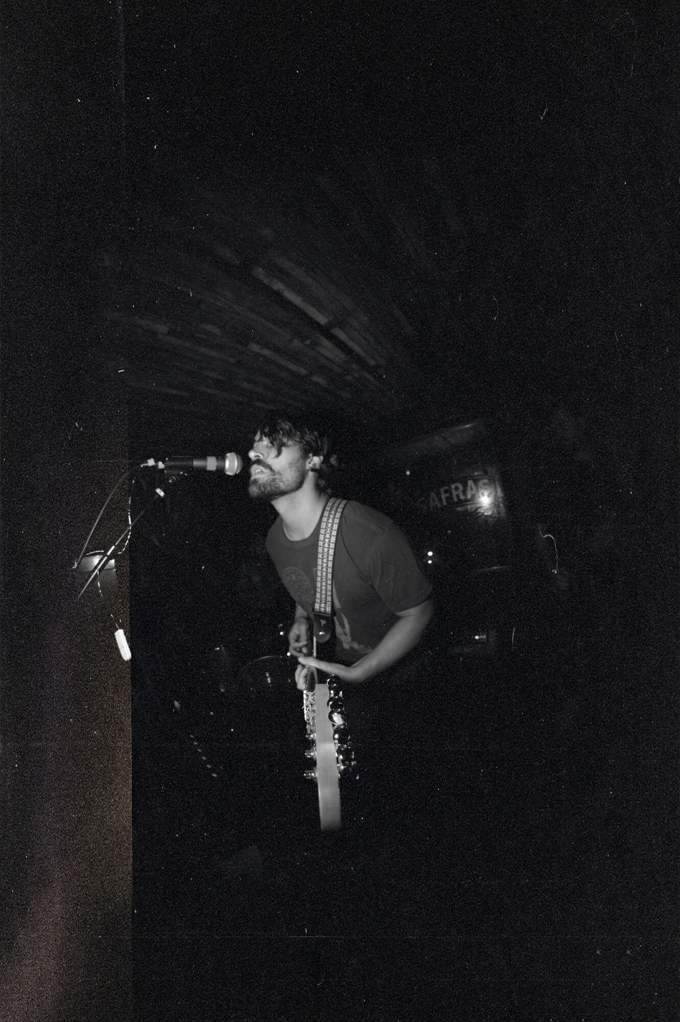 Person singing into a microphone on stage with dark background, holding a guitar, illuminated by stage lights.