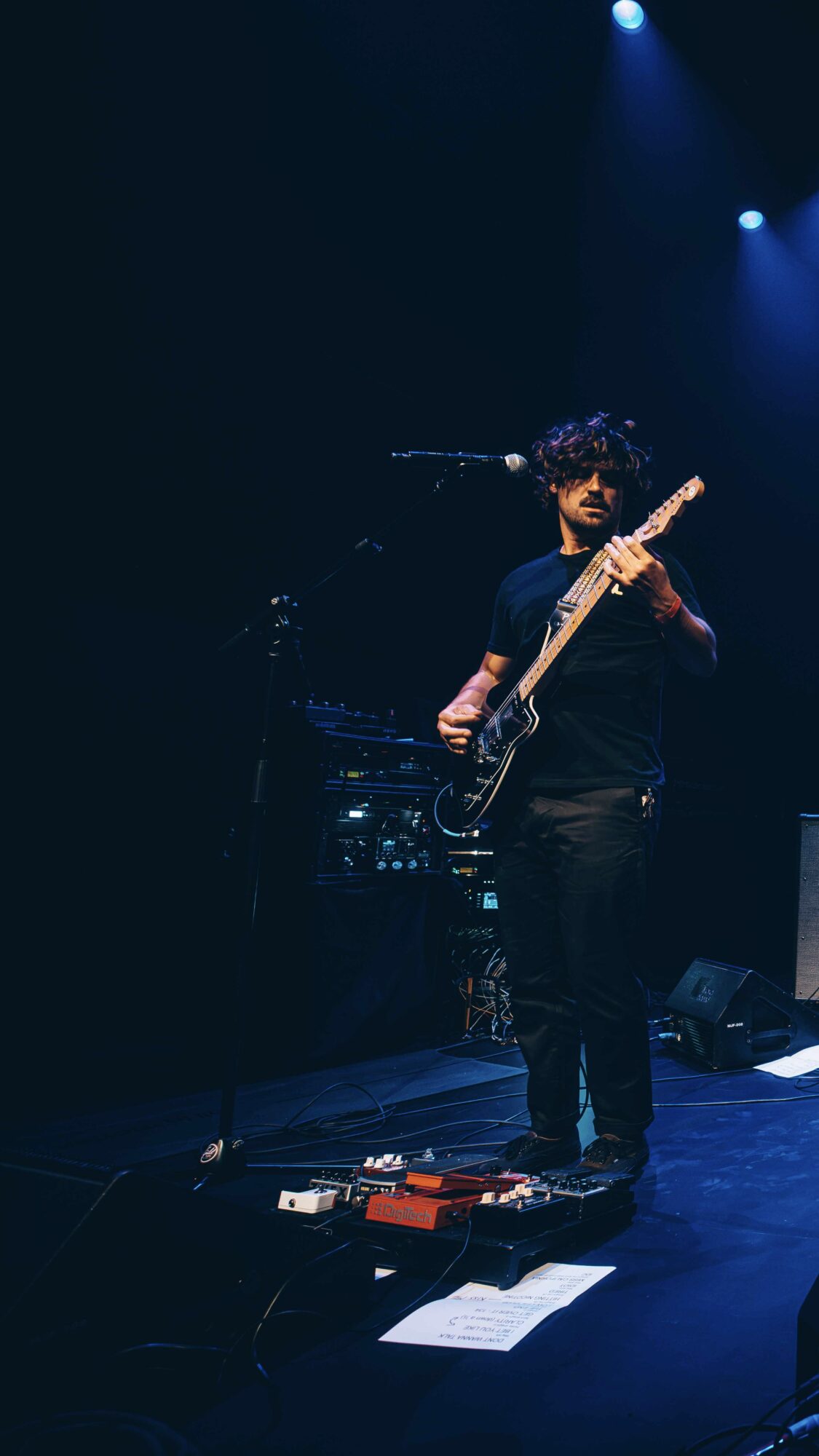 Person playing an electric guitar on stage with blue lighting, standing near equipment and pedals.
