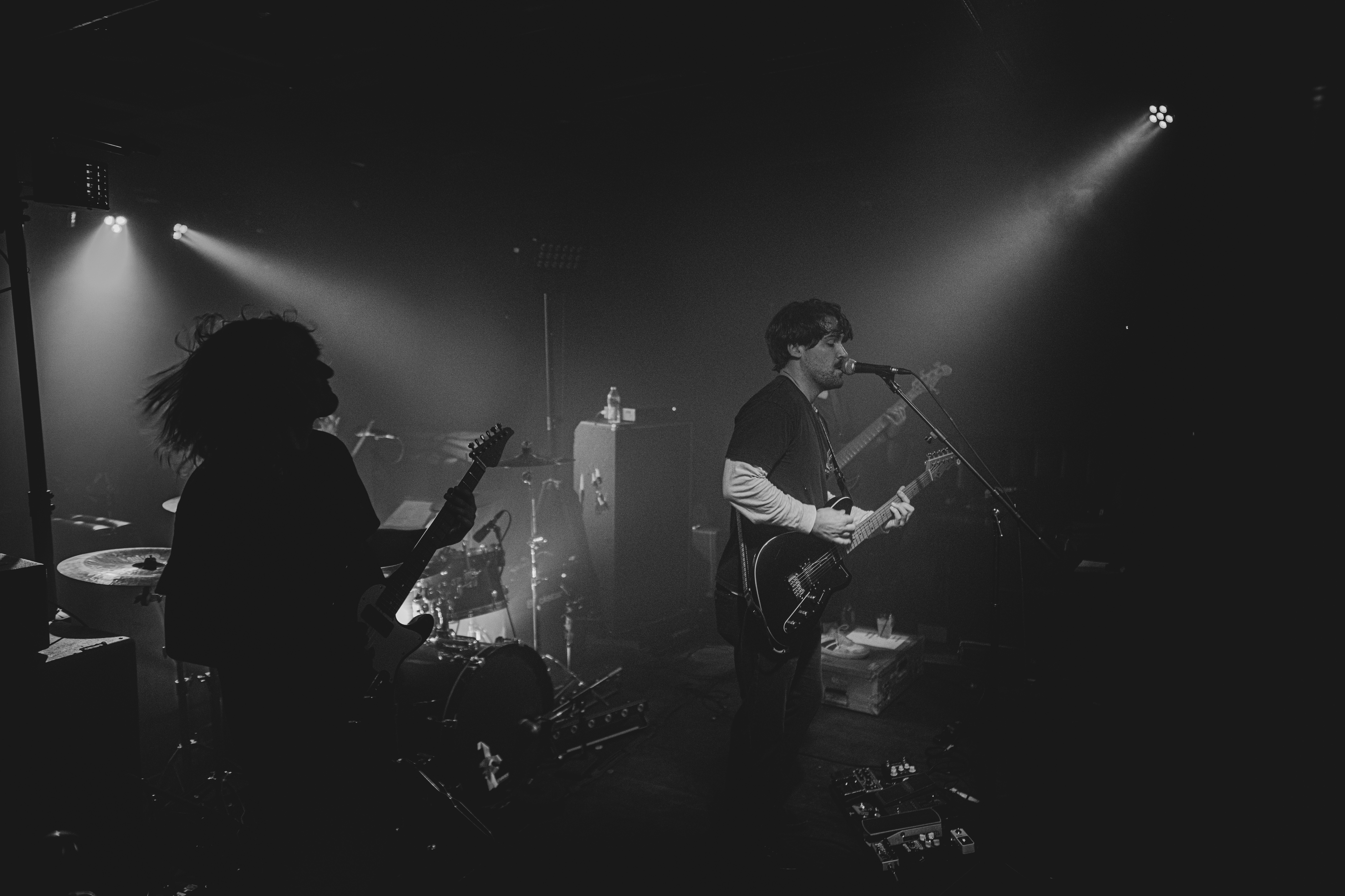 Musicians performing on stage with guitars, microphone, and stage lighting, in black and white.