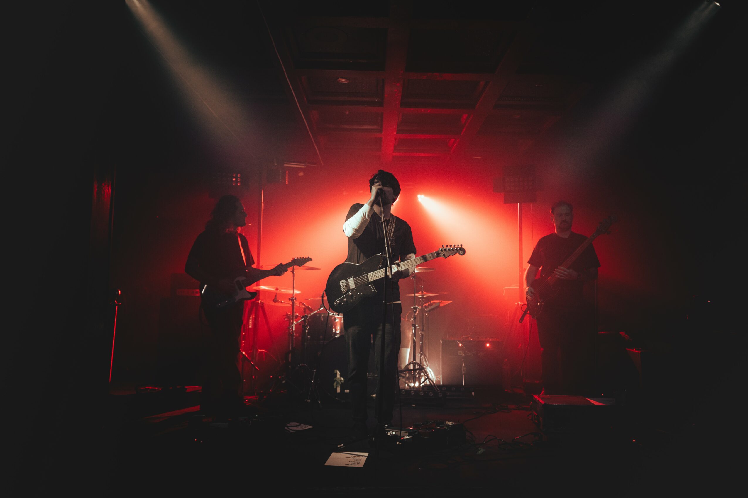 Band performing on stage with red backlighting, four musicians playing instruments, one singing into a microphone, dark atmosphere