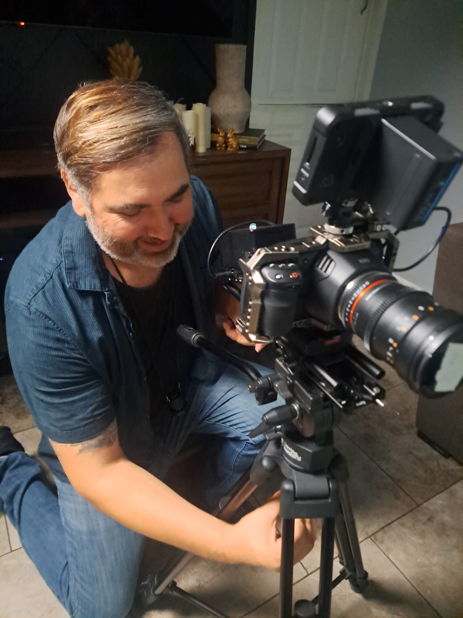 Man operating a professional camera on a tripod indoors, with a monitor attached, in a room with furniture and decor.