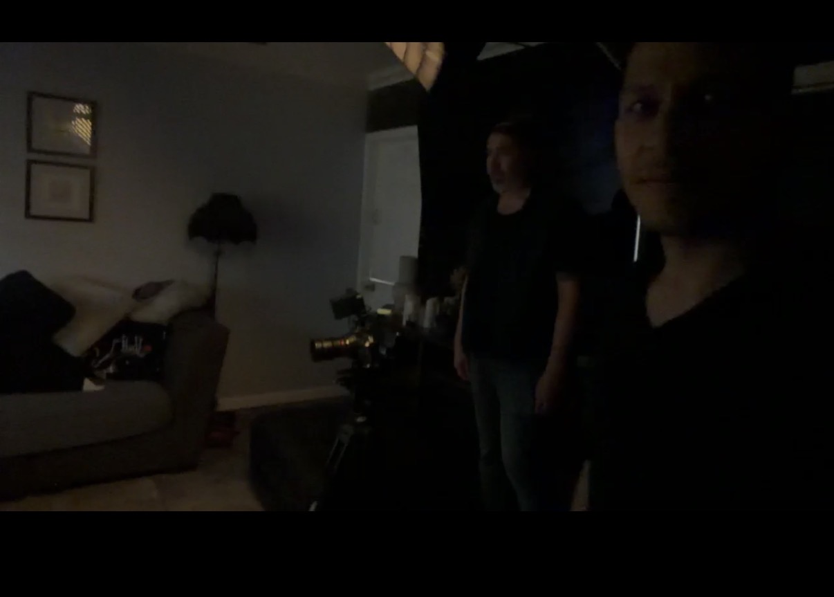 Two people stand in a dark room with a sofa, lamp, and framed pictures on the wall, faintly visible.