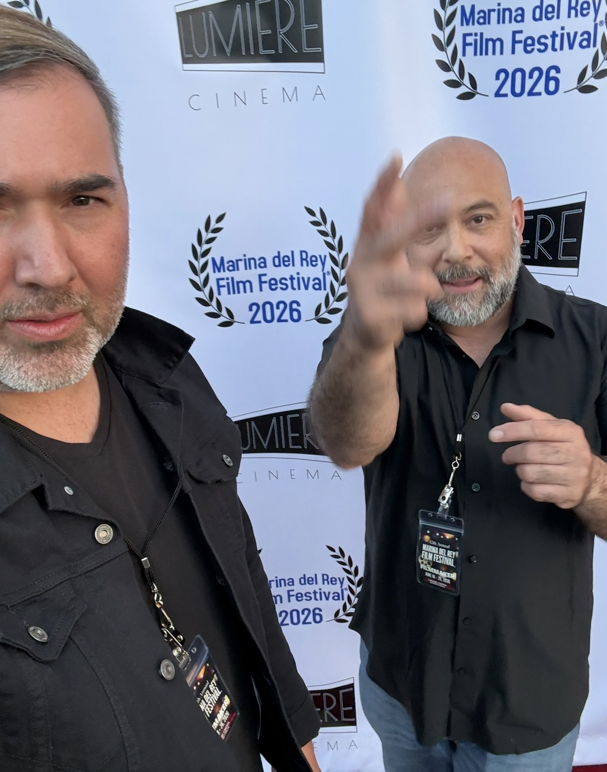 Two men standing in front of a backdrop with festival logos, one smiling and waving, the other partially visible.