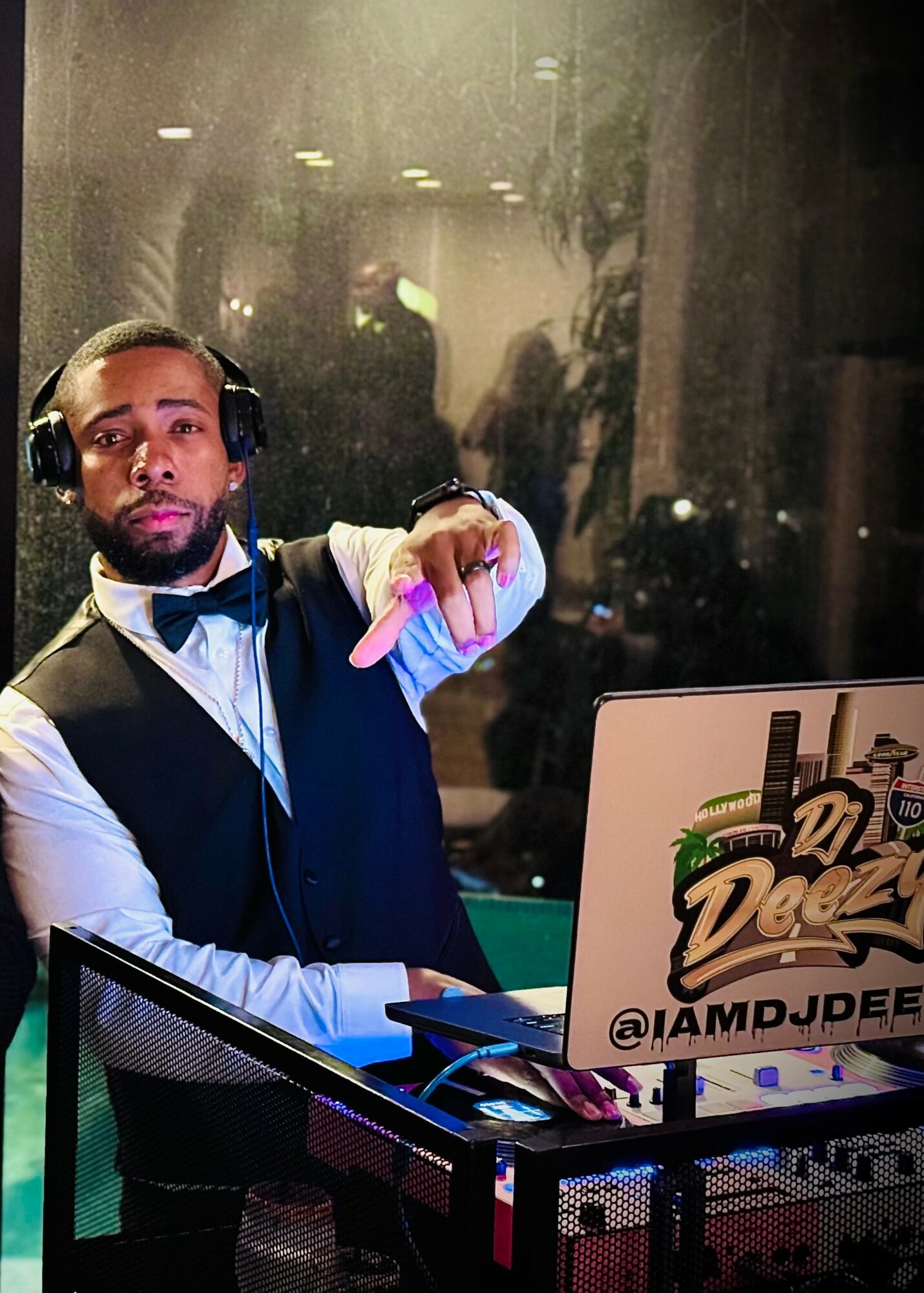 DJ wearing headphones and a vest, pointing at the camera, with a laptop displaying cityscape and palm trees, in a dimly lit room.