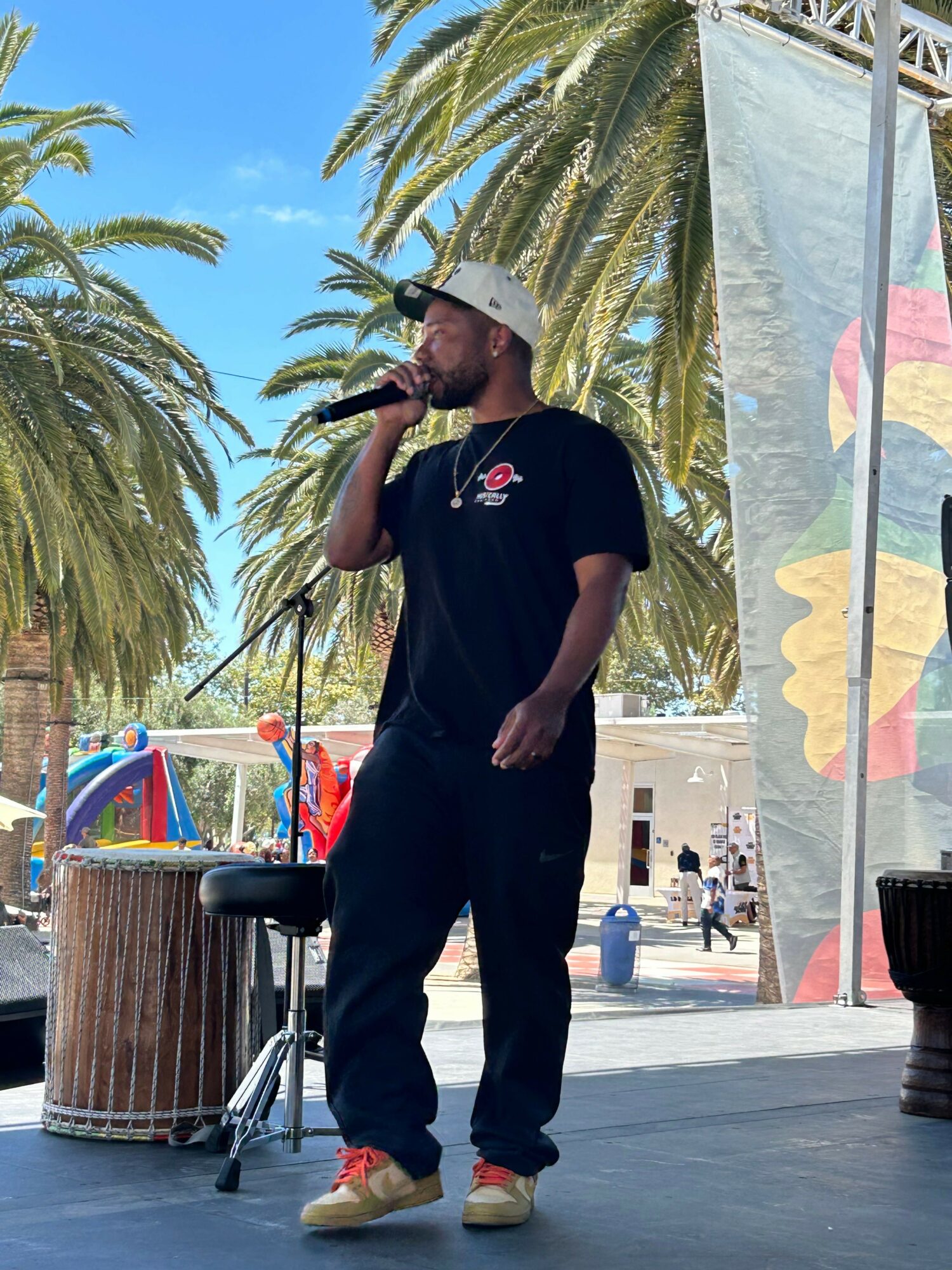 Person standing outdoors near palm trees, wearing black shirt and pants, with a colorful umbrella and trash bin nearby.