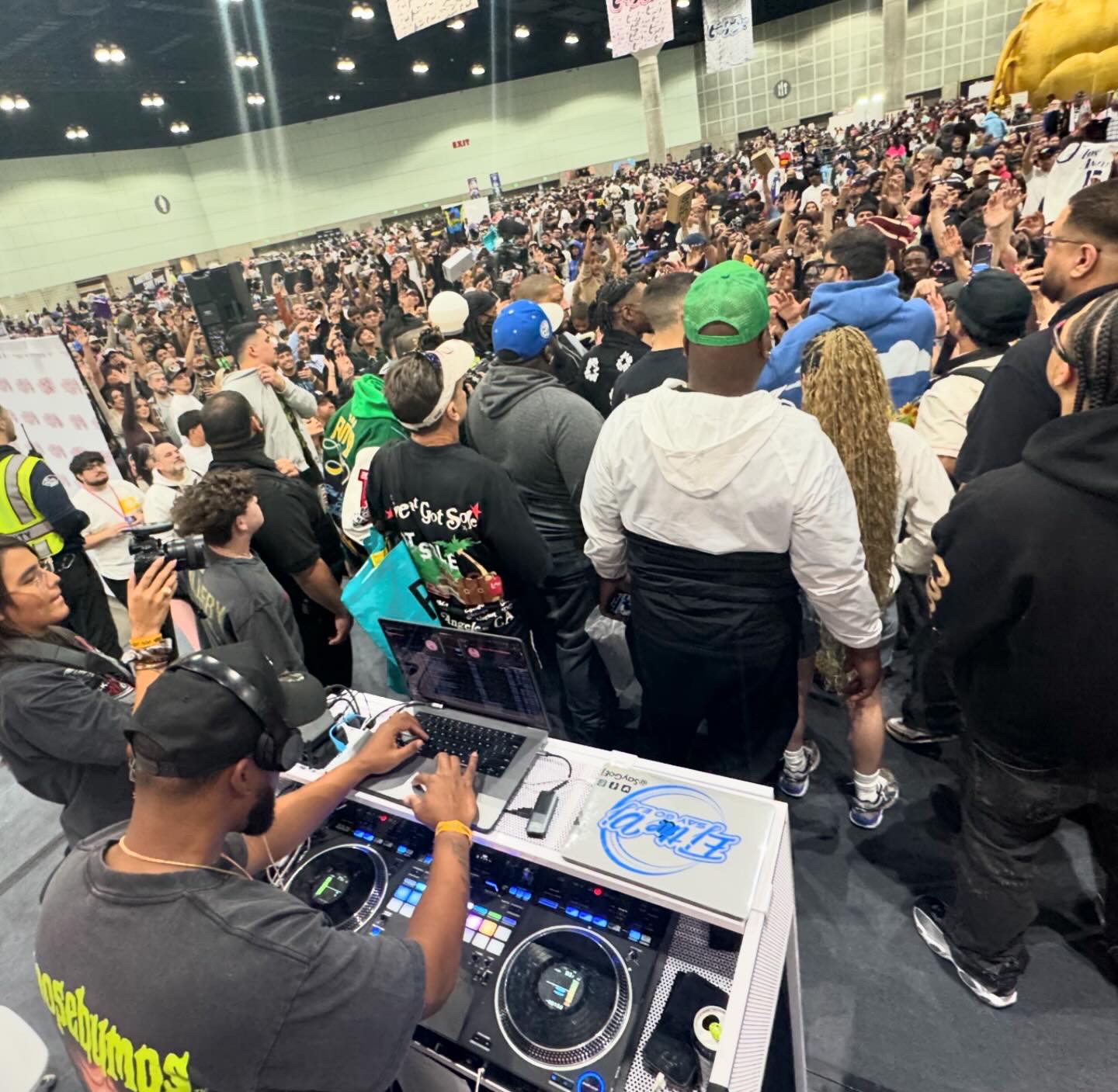 Crowd gathered at an indoor event with a DJ playing music in the foreground, people standing and watching the scene.