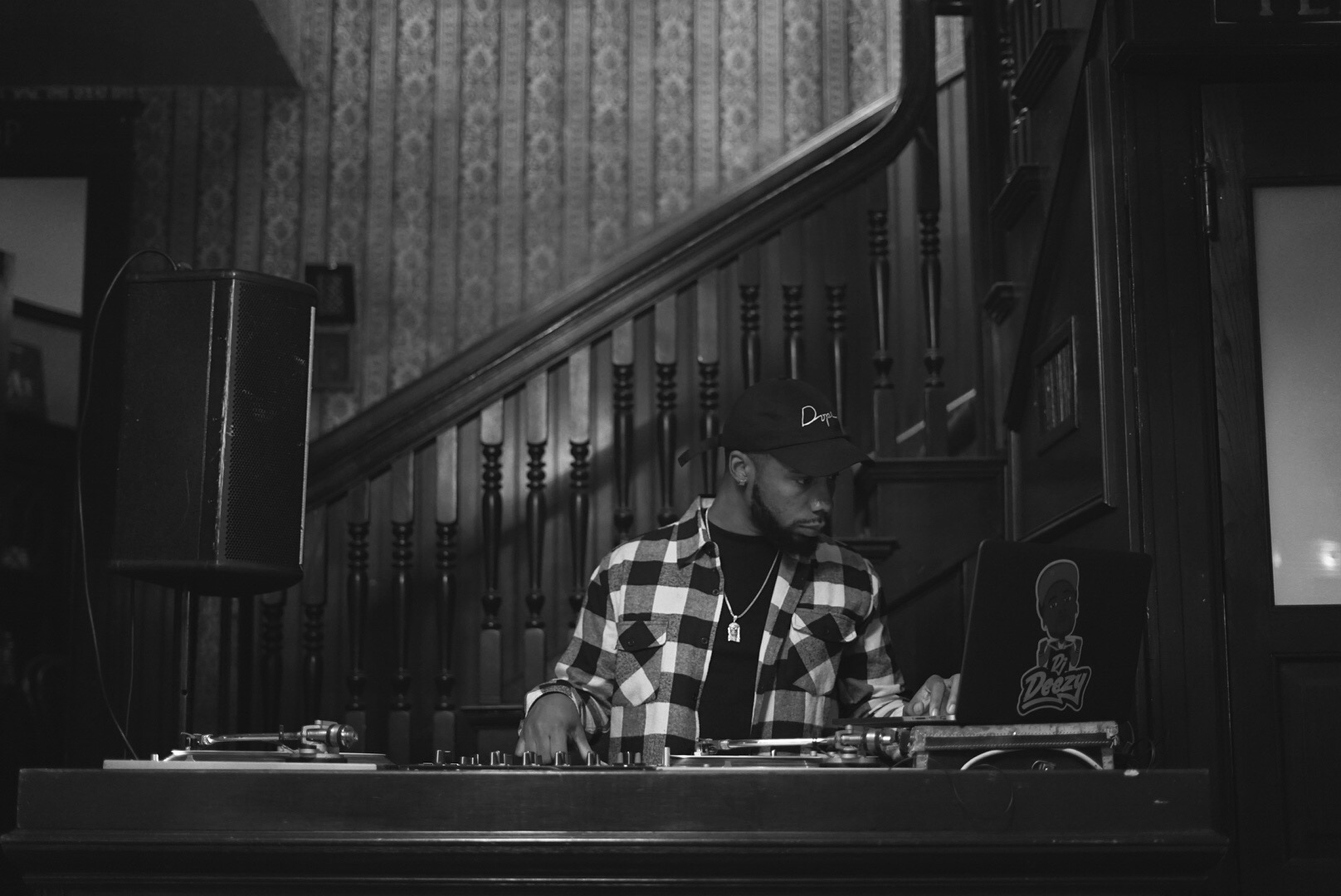 Person wearing a checkered shirt and cap, working with DJ equipment in a dimly lit room with wooden stairs in background.