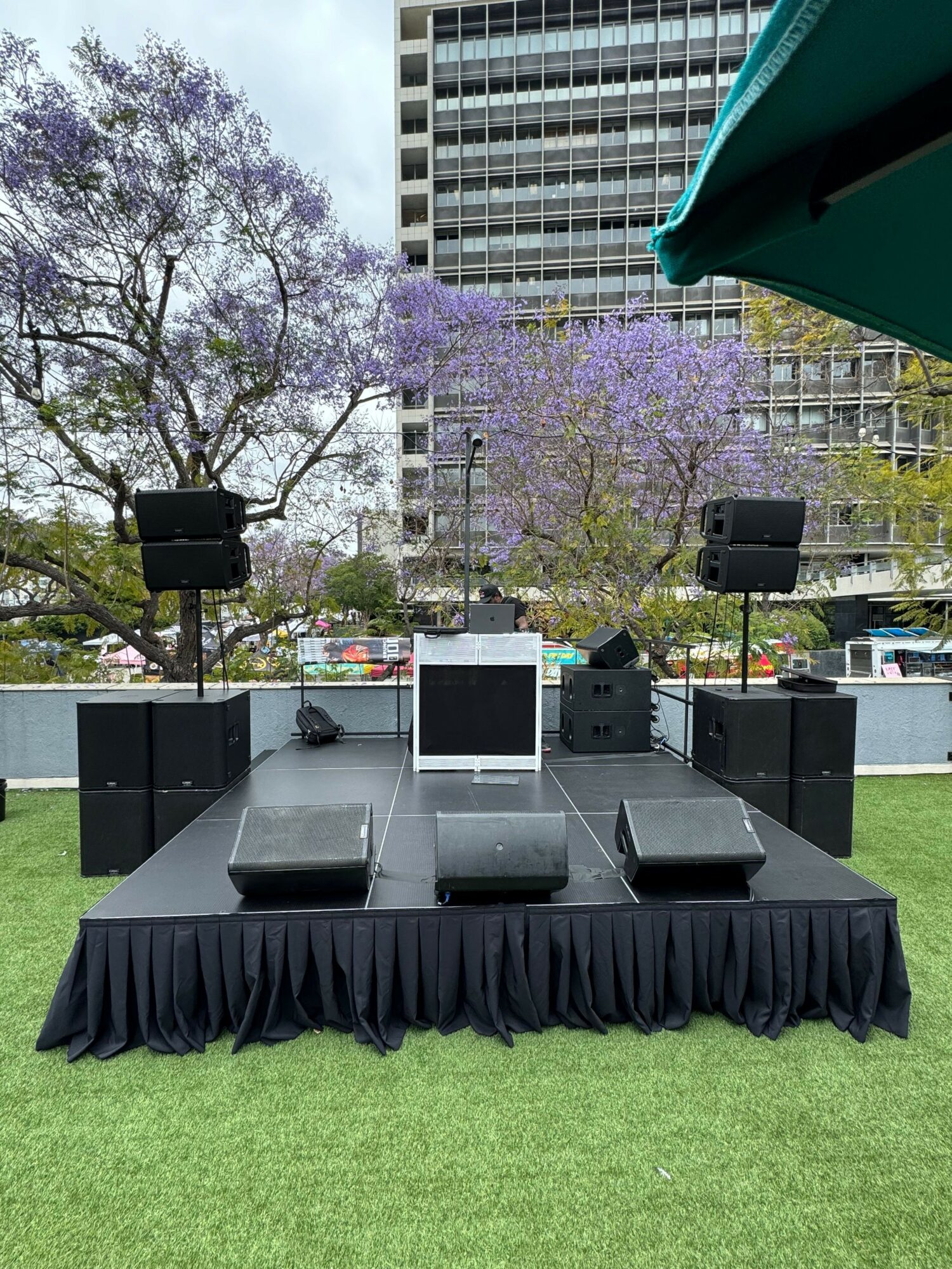 Outdoor stage setup with speakers, monitors, and a table with a black cloth, on a grassy area with trees and buildings.