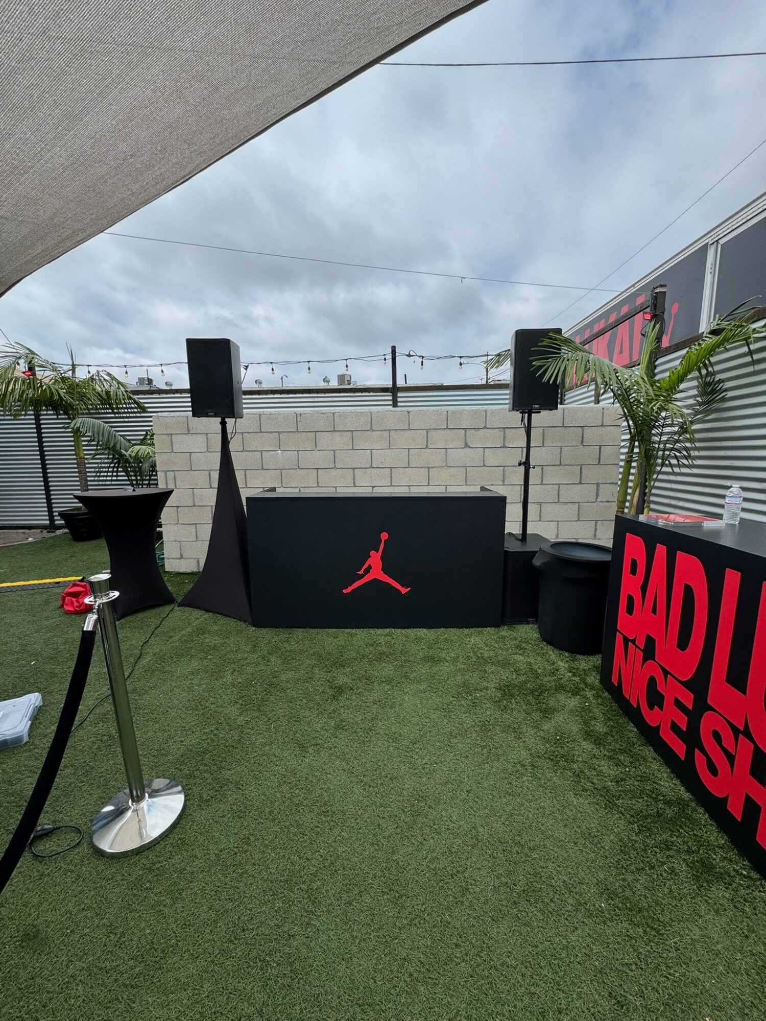 Outdoor event setup with speakers, a DJ booth, and a red sign reading 'RADIO MCS'. Green grass and potted plants are visible.