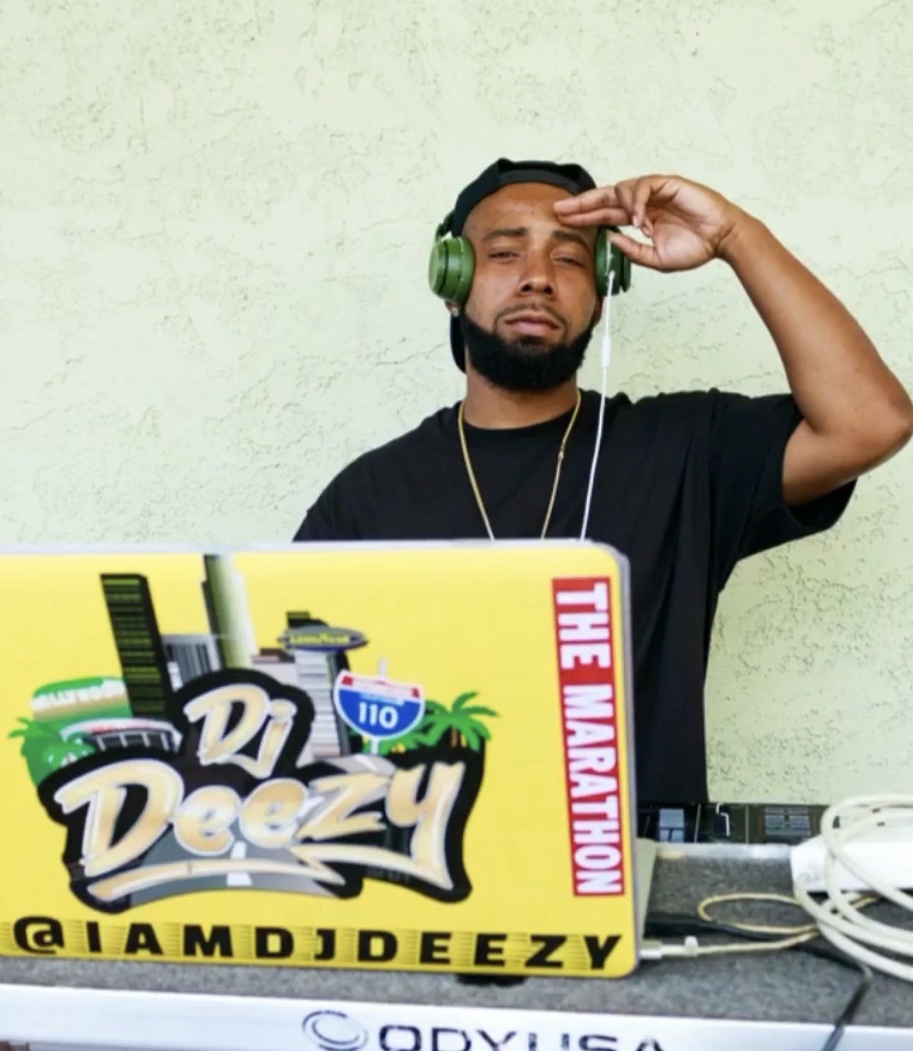 Man with headphones and a beard, standing behind a DJ setup, saluting with his right hand, against a light green wall.
