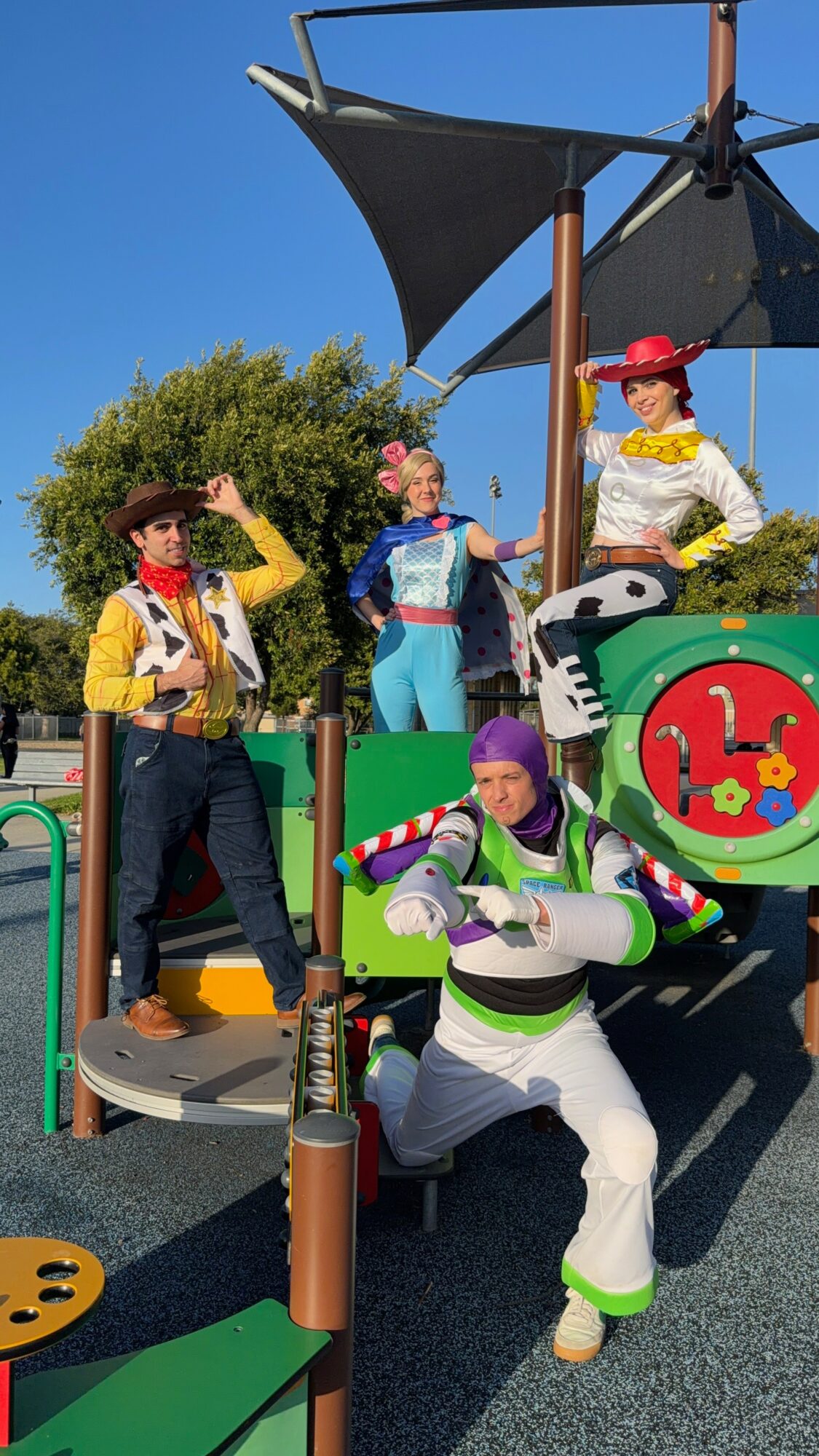 Four people dressed as Toy Story characters pose outdoors with a playground and blue sky, some wearing hats and costumes.