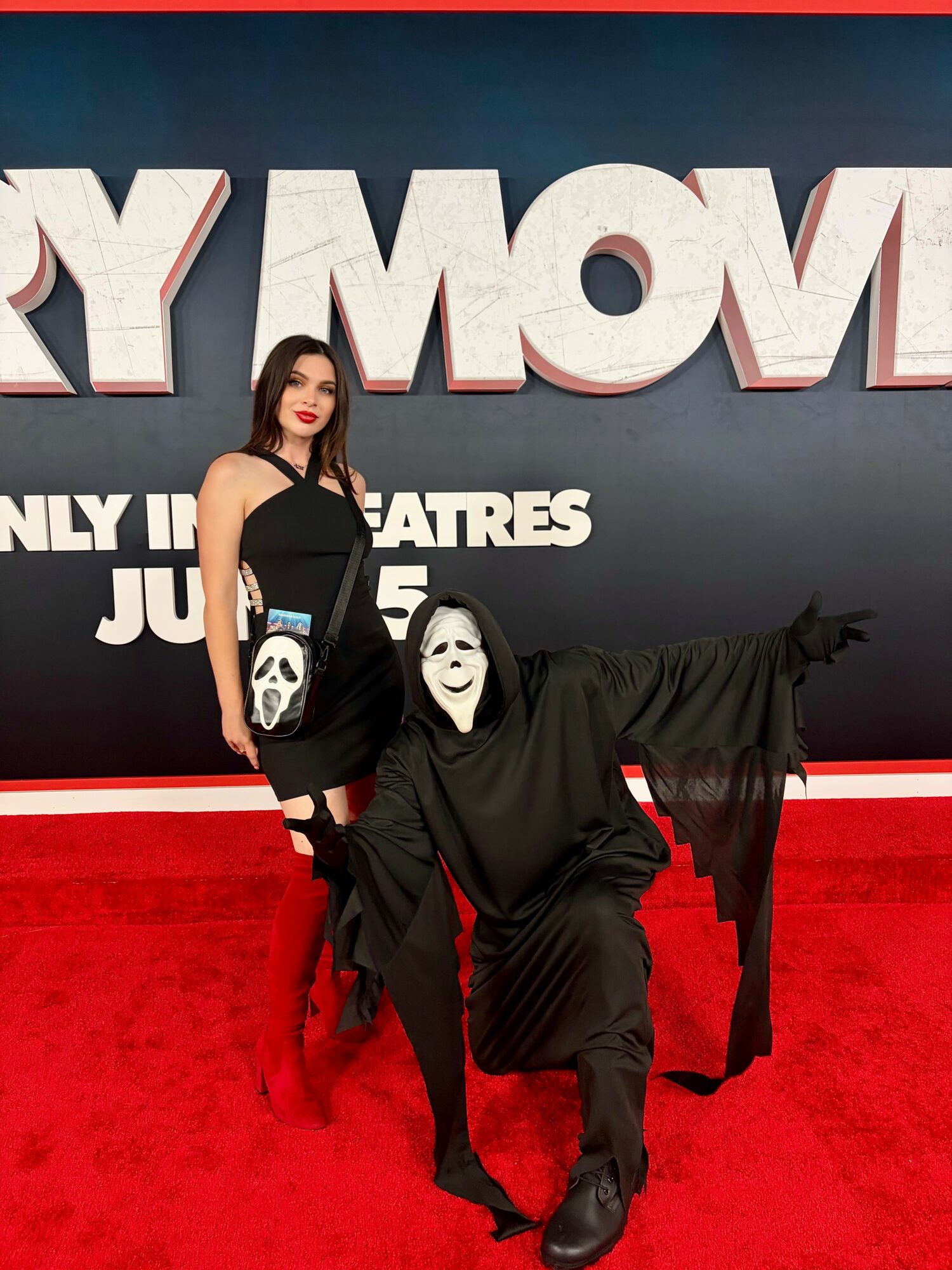 A woman in a black dress and a person in a black costume with a white mask pose on a red carpet in front of a large movie sign.