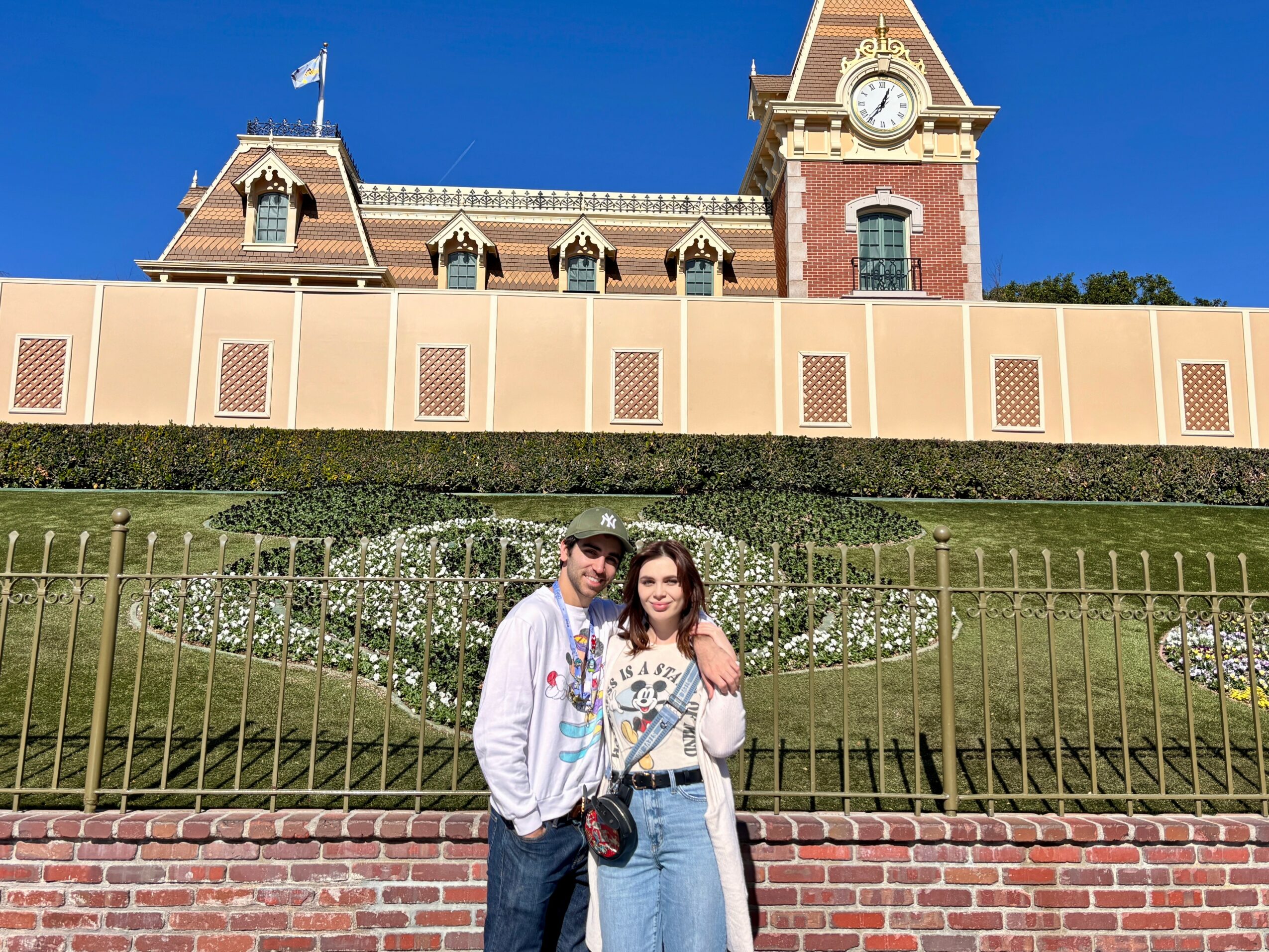Two people stand in front of a castle-like building with a clock tower, fence, and landscaped garden, sunny day.