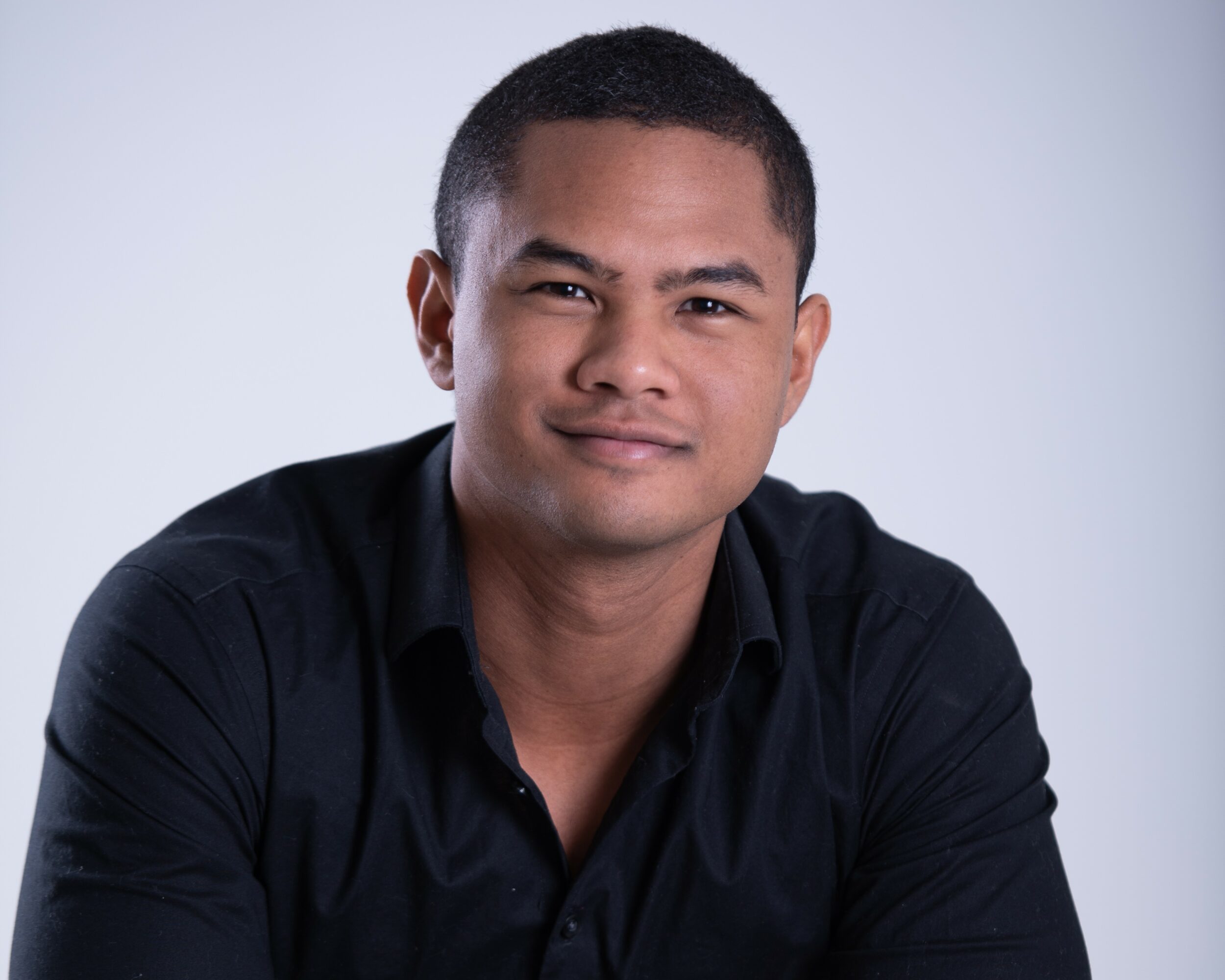 Portrait of a young man with short hair, wearing a black shirt, smiling slightly against a light background.