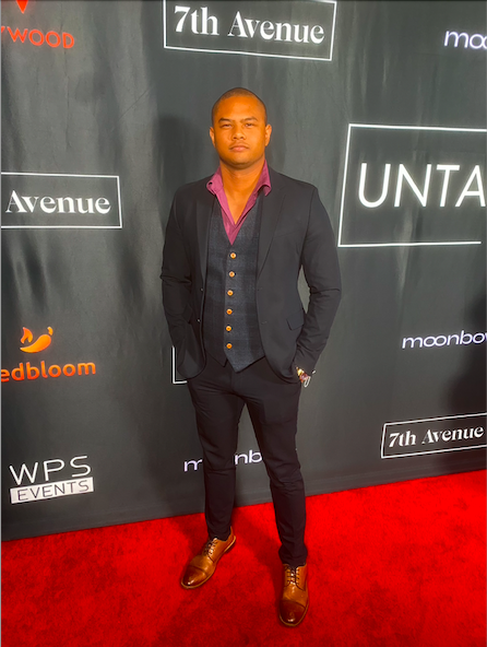 Man in a suit standing on red carpet in front of a black backdrop with text and logos.