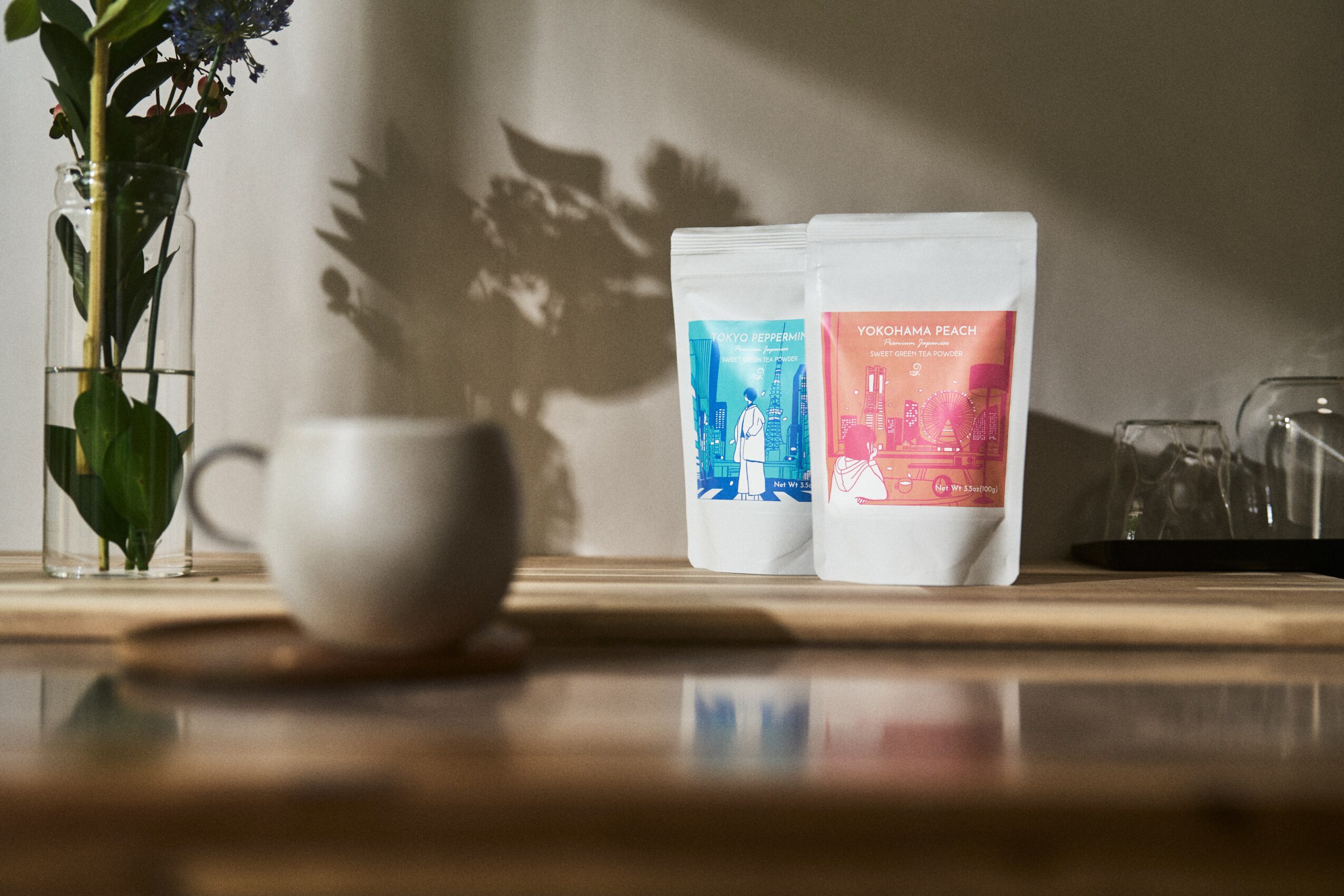 Two packages of coffee or tea on a wooden surface with a blurred background, a white mug, and a vase with flowers.