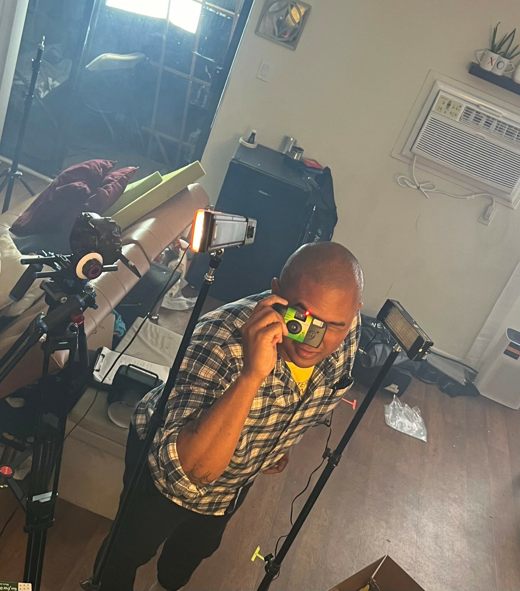 Person taking a selfie with a camera, wearing glasses, in a room with cameras and equipment, and a window with sunlight.